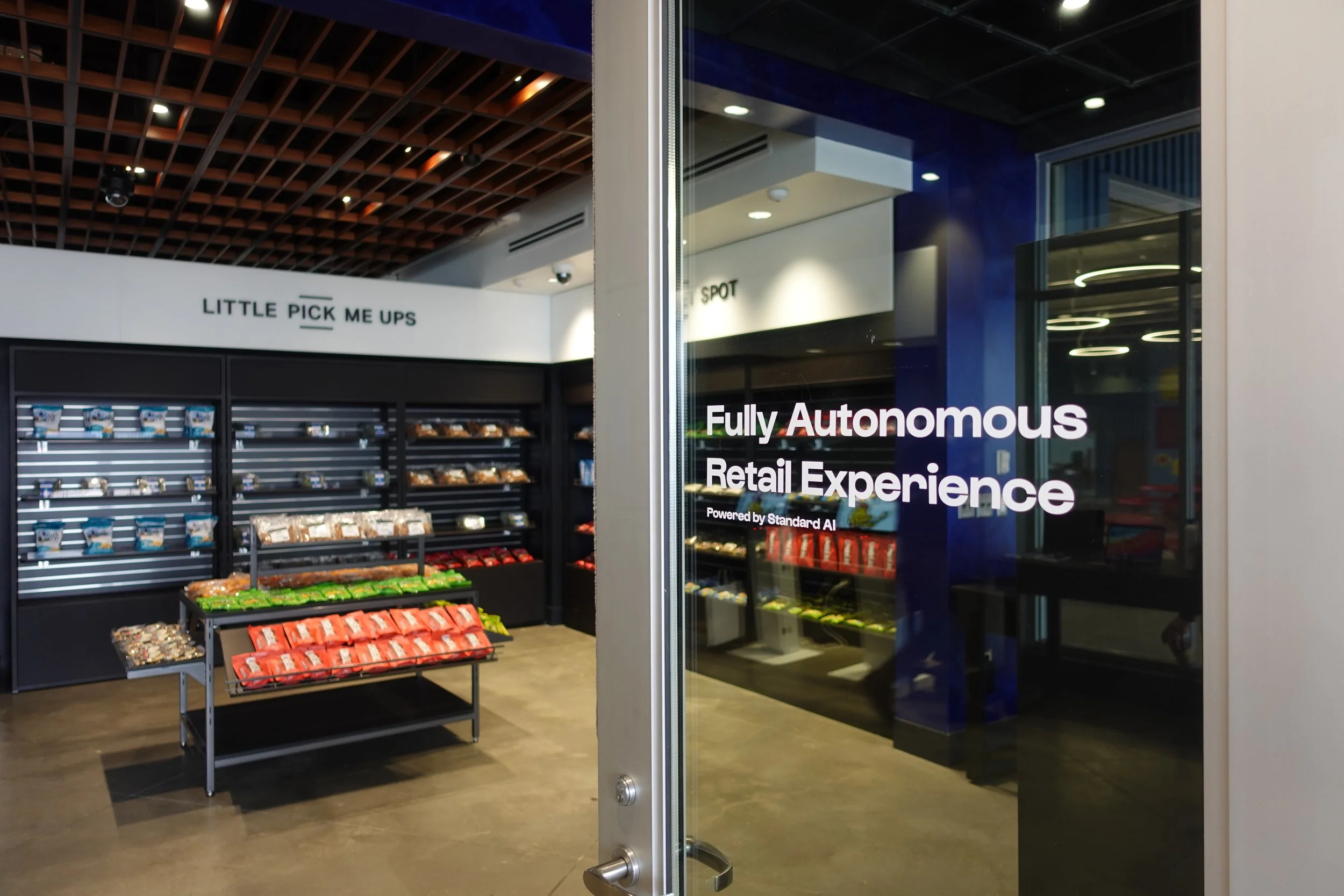 Interior of a store with shelves of snacks and a sign that reads 'Little Pick Me Ups'. A glass door with a sign stating 'Fully Autonomous Retail Experience' powered by Standard AI.