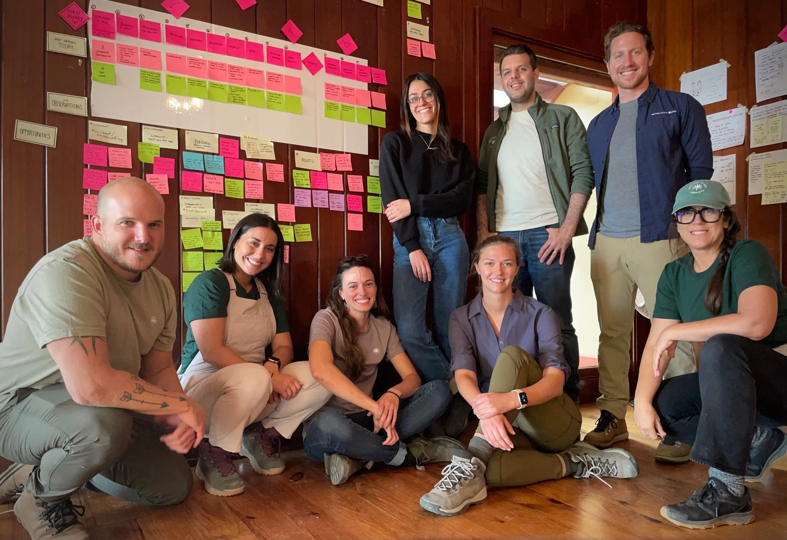 A group of nine diverse young adults posing for a photo in a wooden room with a large whiteboard covered in colorful sticky notes and notes on the wall. They are smiling, sitting and standing casually.