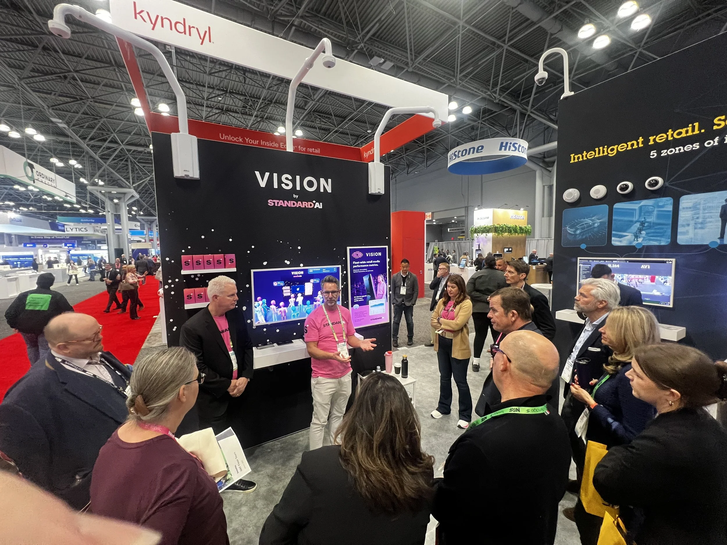 A busy trade show booth with a black display board titled 'VISION by STANDARD AI,' featuring digital screens and informational posters. Several people are gathered around, listening to a presenter, with booths and attendees visible in the background. Hanging signs read 'kyndryl' and 'HiStone,' and the exhibit area is bustling with activity under an industrial-style ceiling.