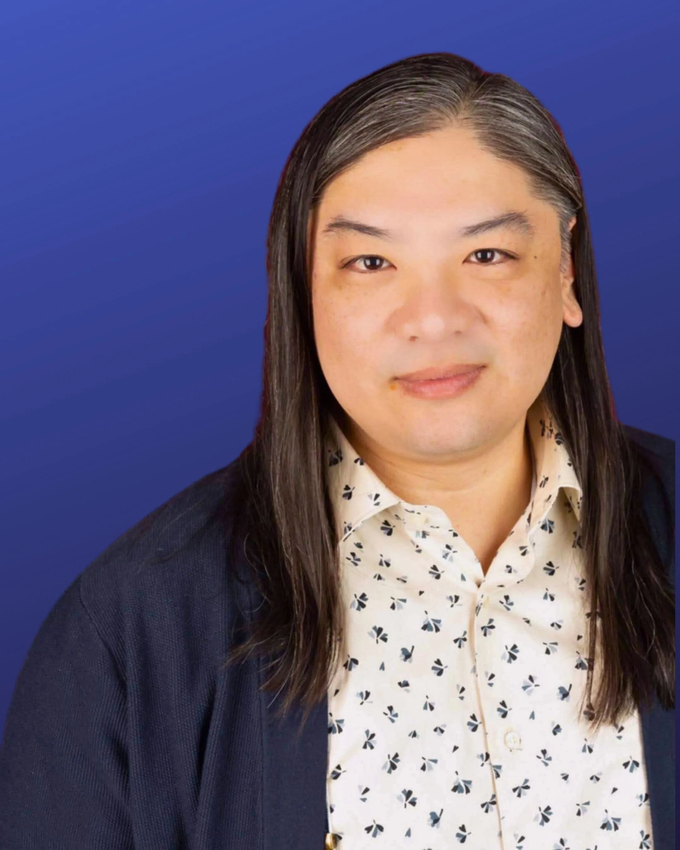 Person with long hair, wearing a patterned shirt and dark sweater, against a blue background.