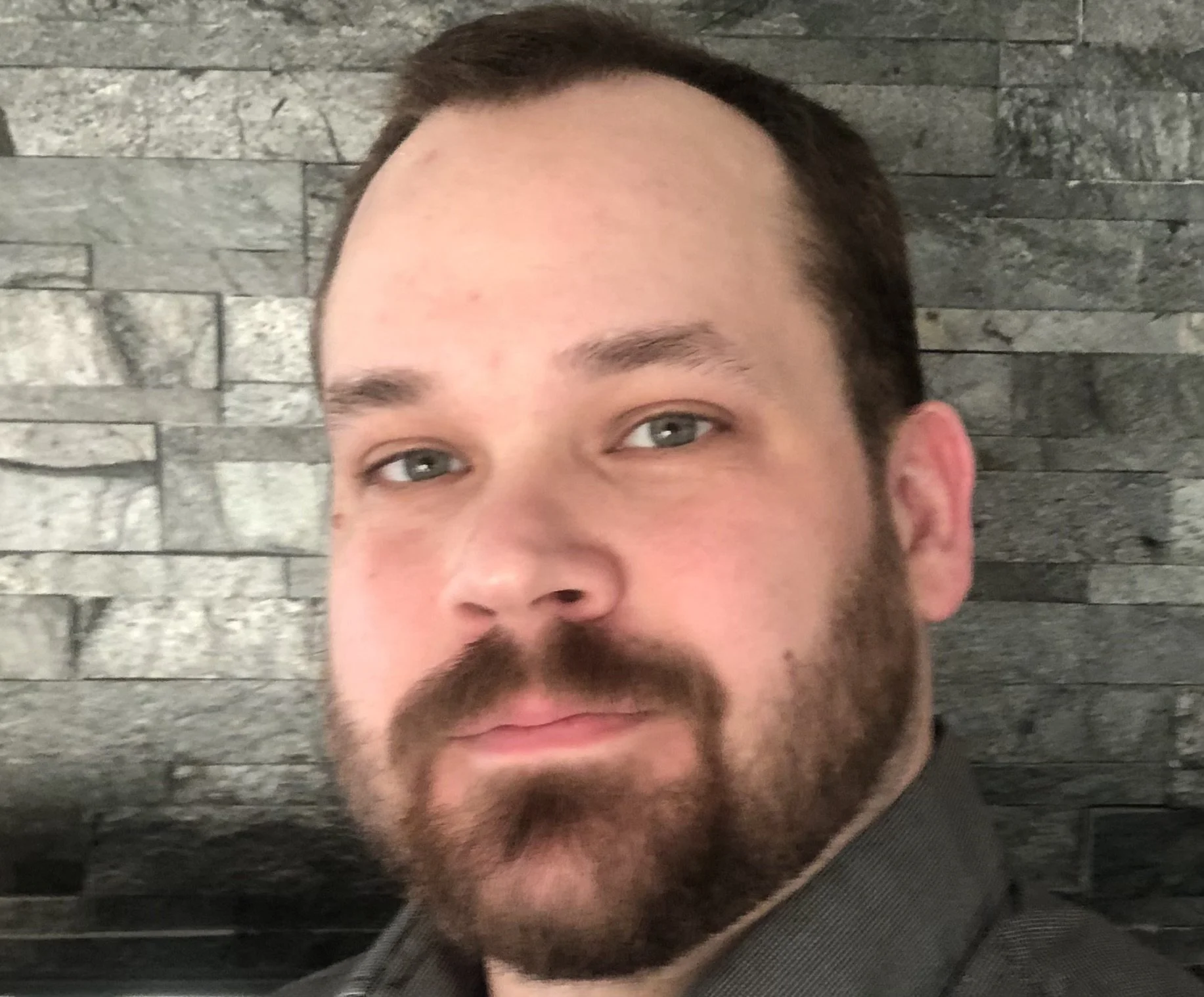 Close-up of a person with a beard, wearing a gray shirt, standing in front of a gray stone wall.