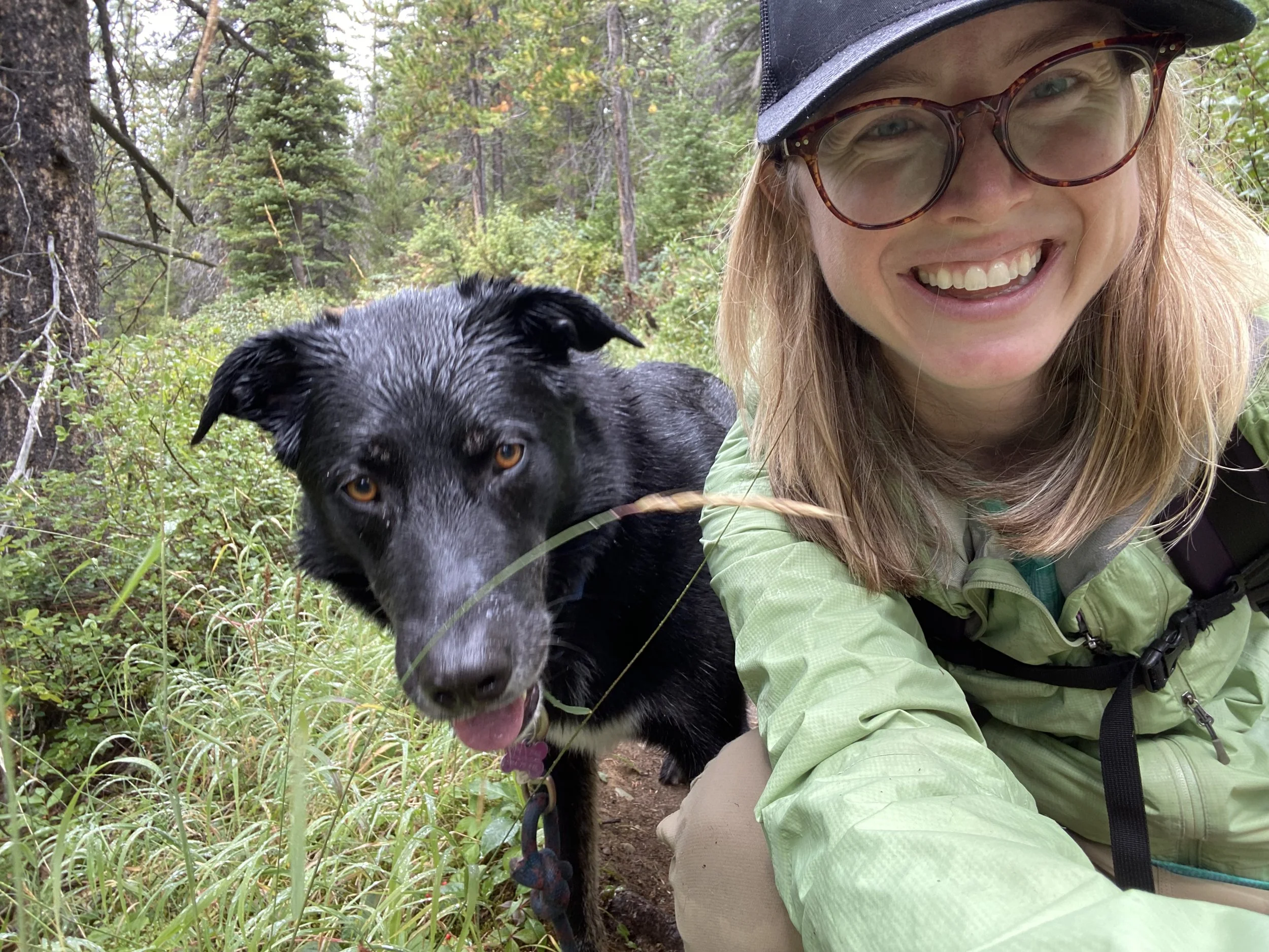 Woman in glasses and a hat smiling with a black dog in a forest.