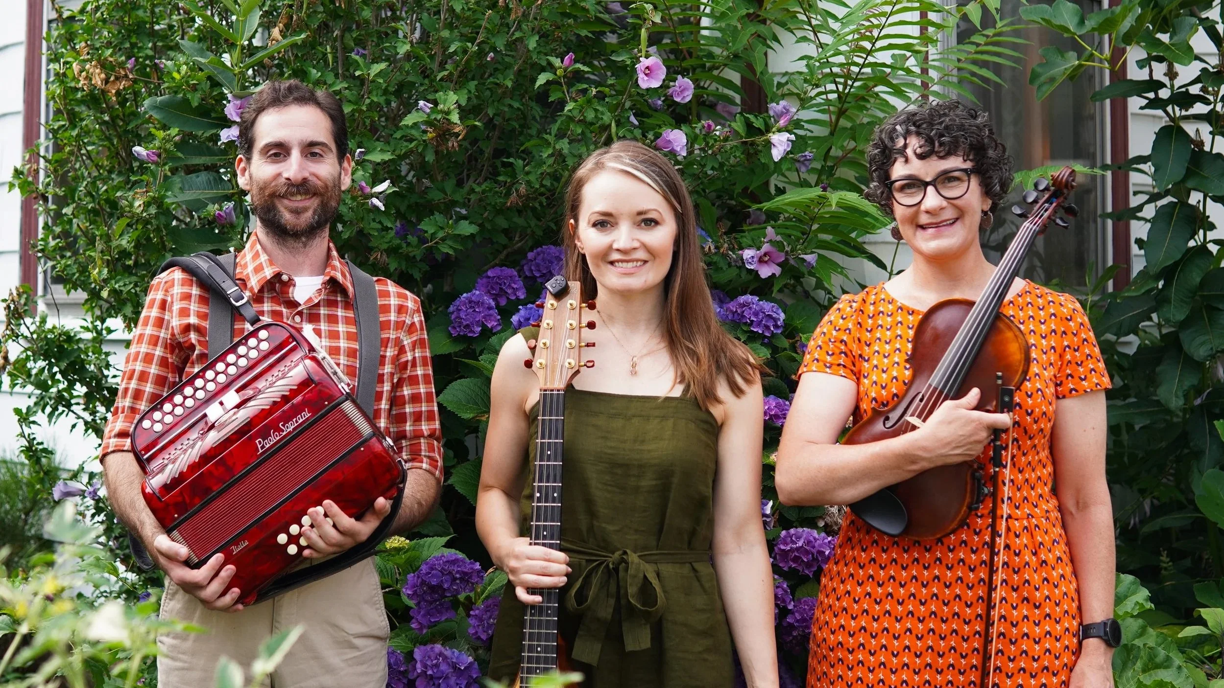 Three musicians holding instruments standing in front of a bush with purple flowers outside a house.