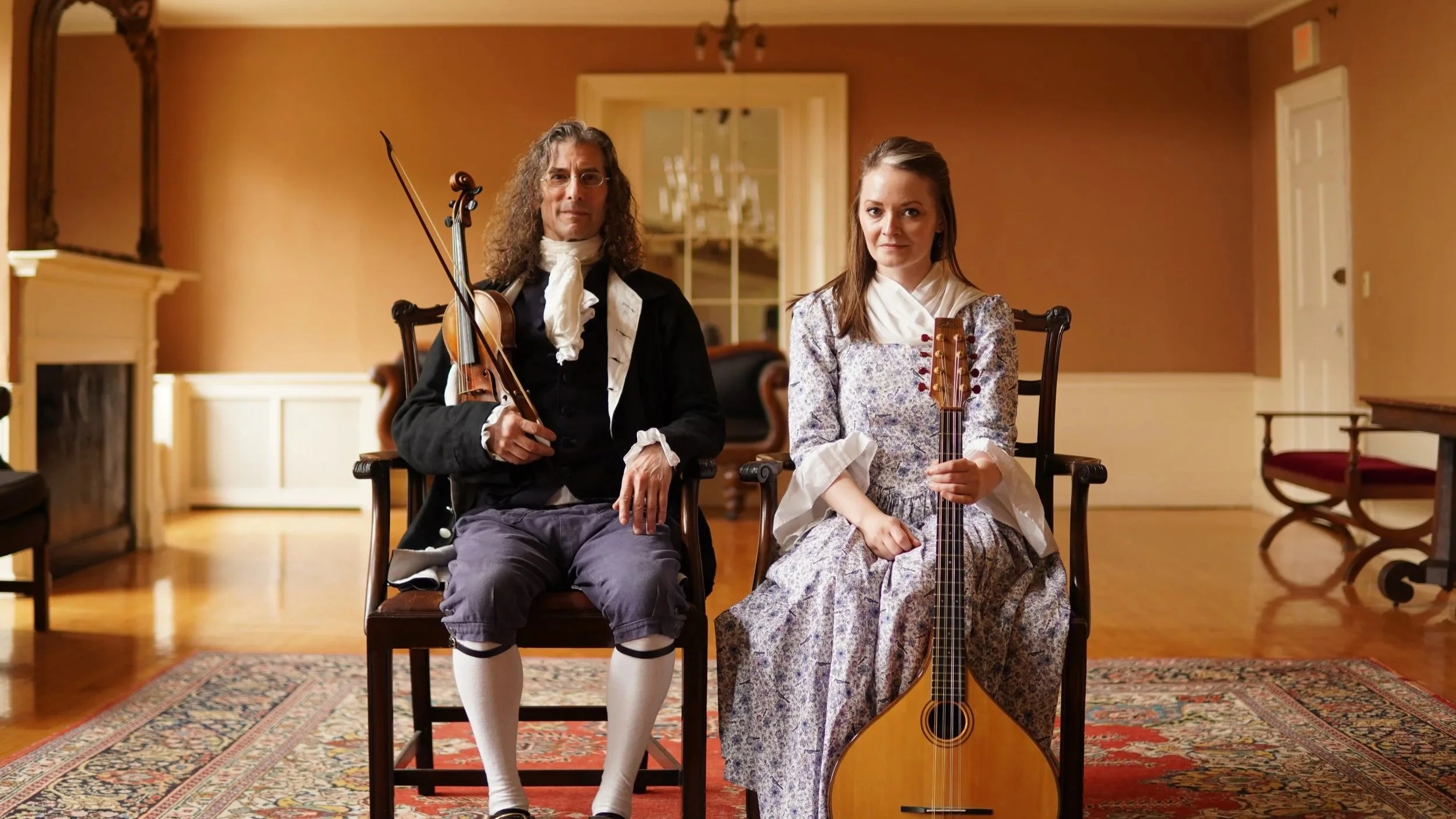Two people dressed in historical clothing sitting in a room, each holding a musical instrument. The man on the left holds a violin, and the woman on the right holds a guitar.