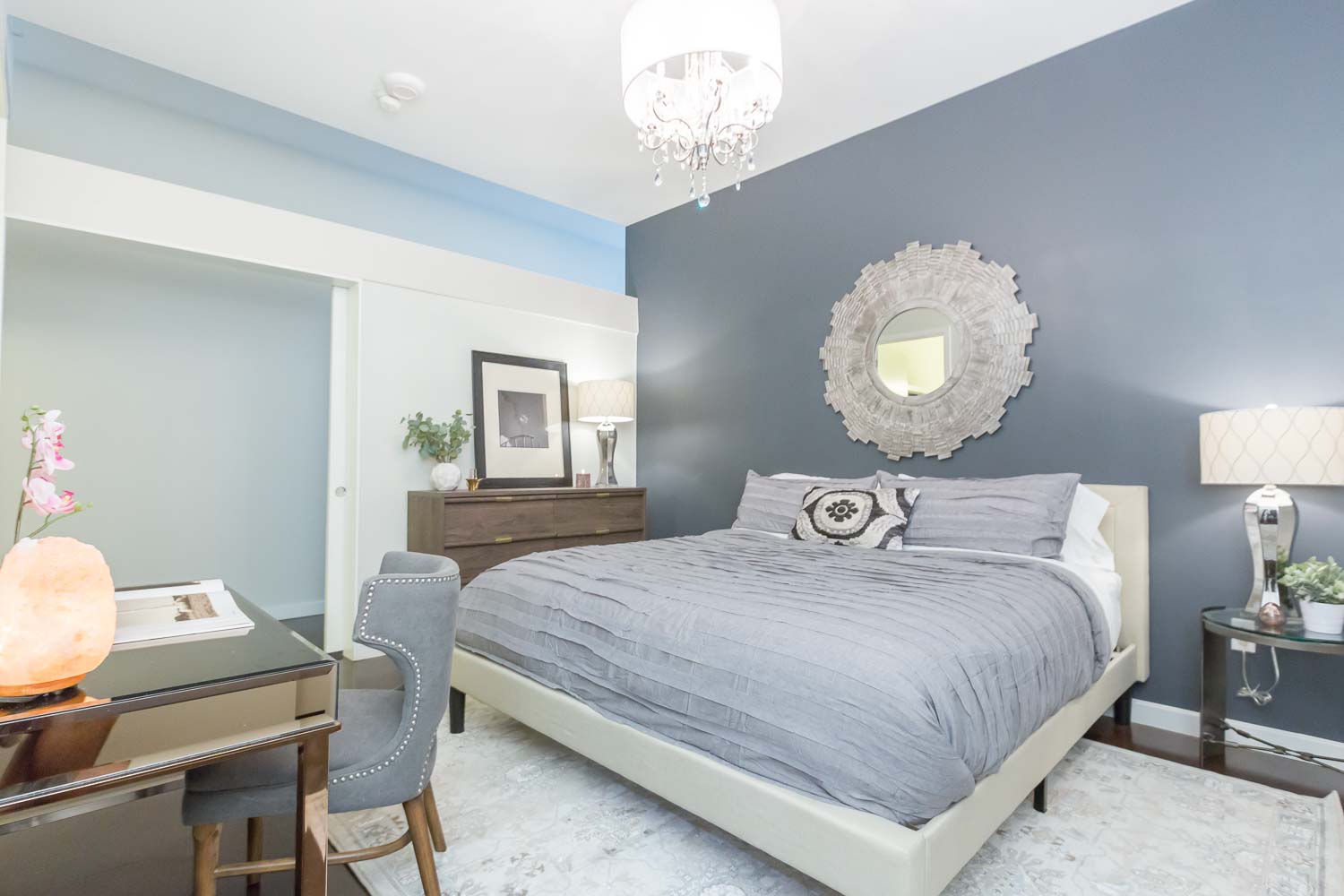 The master bedroom does not have a window, so it was important that we added "depth" and interest. We did this by painting the headboard wall with Sherwin Williams Deep Space. The chandelier adds luxury as does the gray bedding, mirrored console and walnut dresser with gold accents.