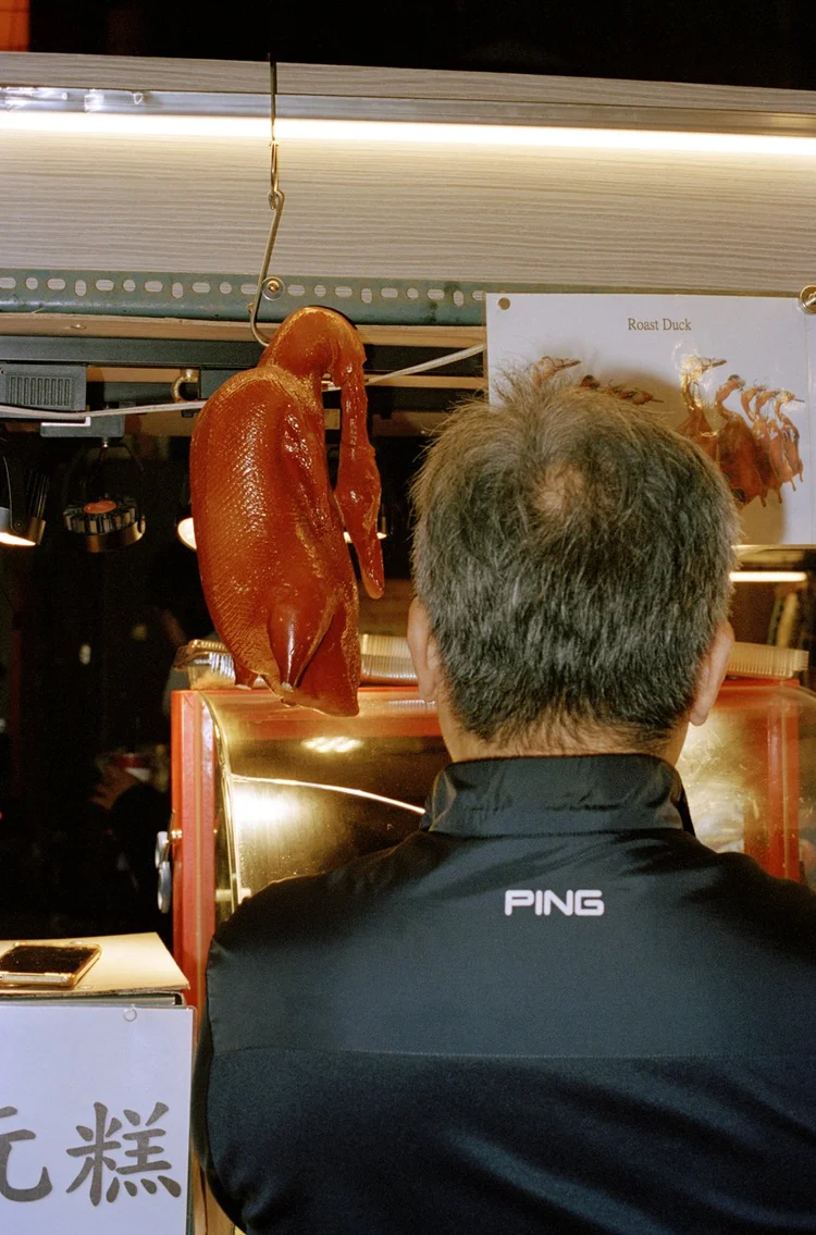 A photograph taken inside a Chinese restaurant or food establishment, showing a person from behind viewing a glossy roasted duck hanging in a display window.