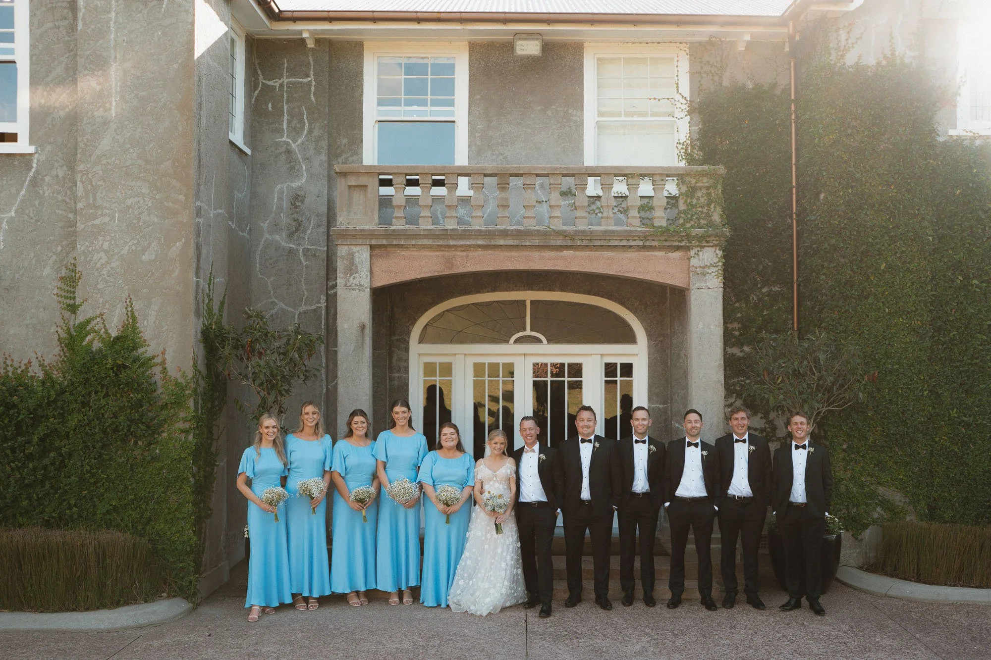 Bridal party standing in front of ivy covered McCallum Residence building with sun flare