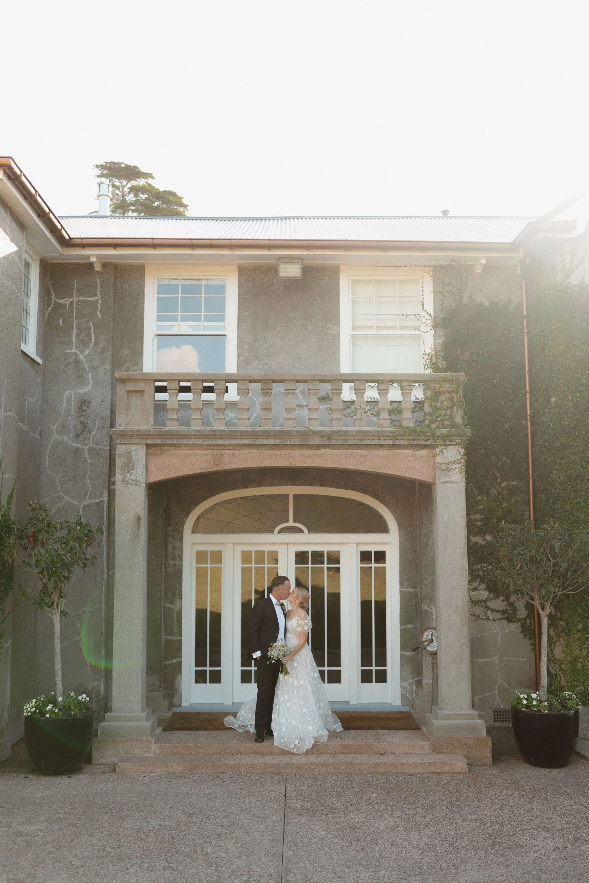 Bride & groom kissing in front of The McCallum Residence ivy covered building sun flaring into shot on Autumn wedding day