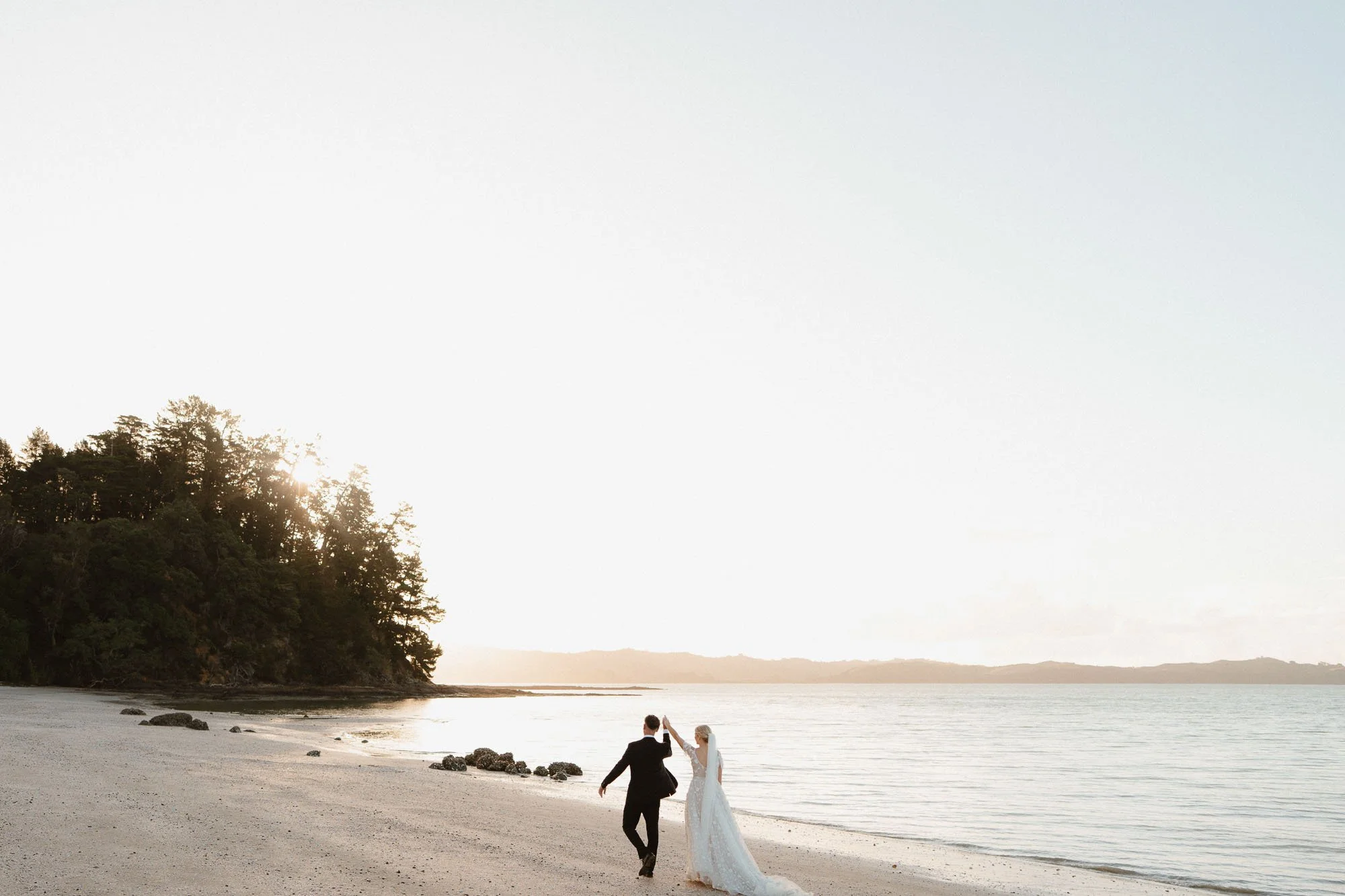 Bride and groom walking along The McCallum Residence beach at dusk, throwing hands in the air in celebration, view of surrounding island in photos