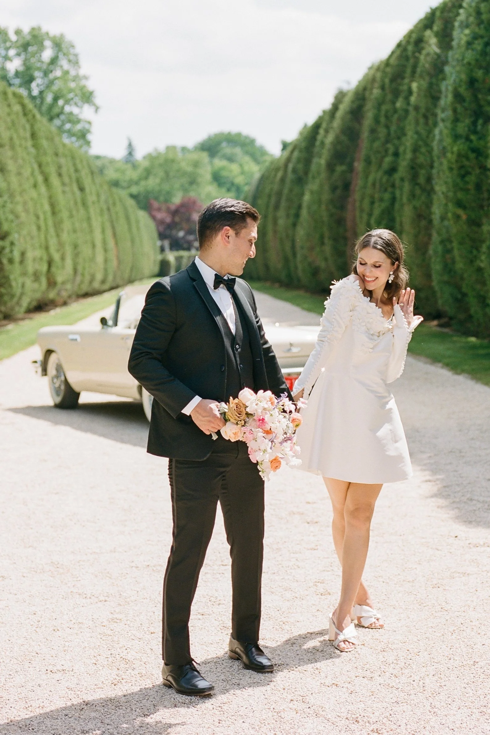 Bride in a short white wedding dress and groom in a black tuxedo sharing a playful moment beside a vintage convertible on a tree-lined estate driveway.