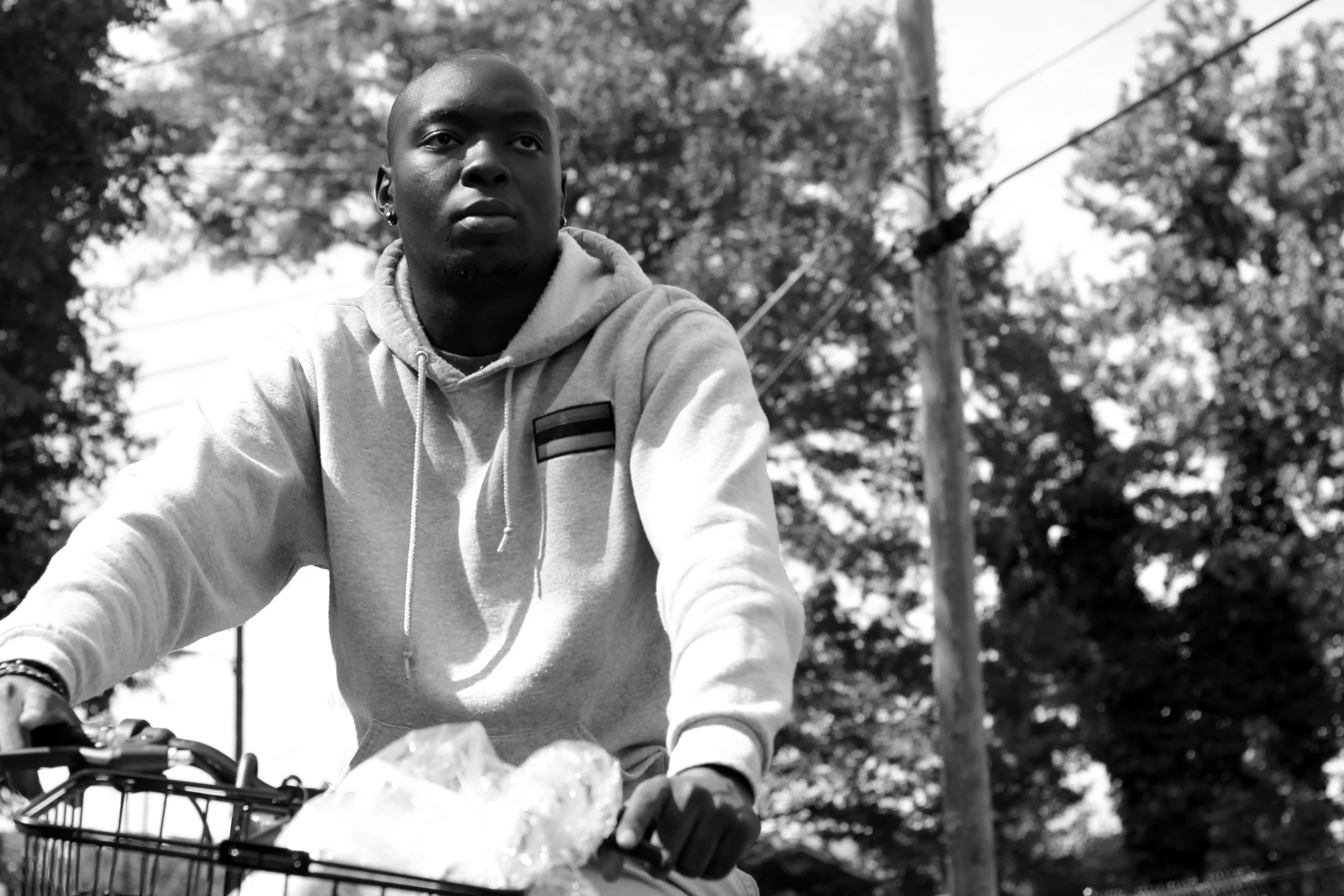 Black and white photo of a young man riding a bicycle outdoors, wearing a hoodie and gloves, with trees and a utility pole in the background.