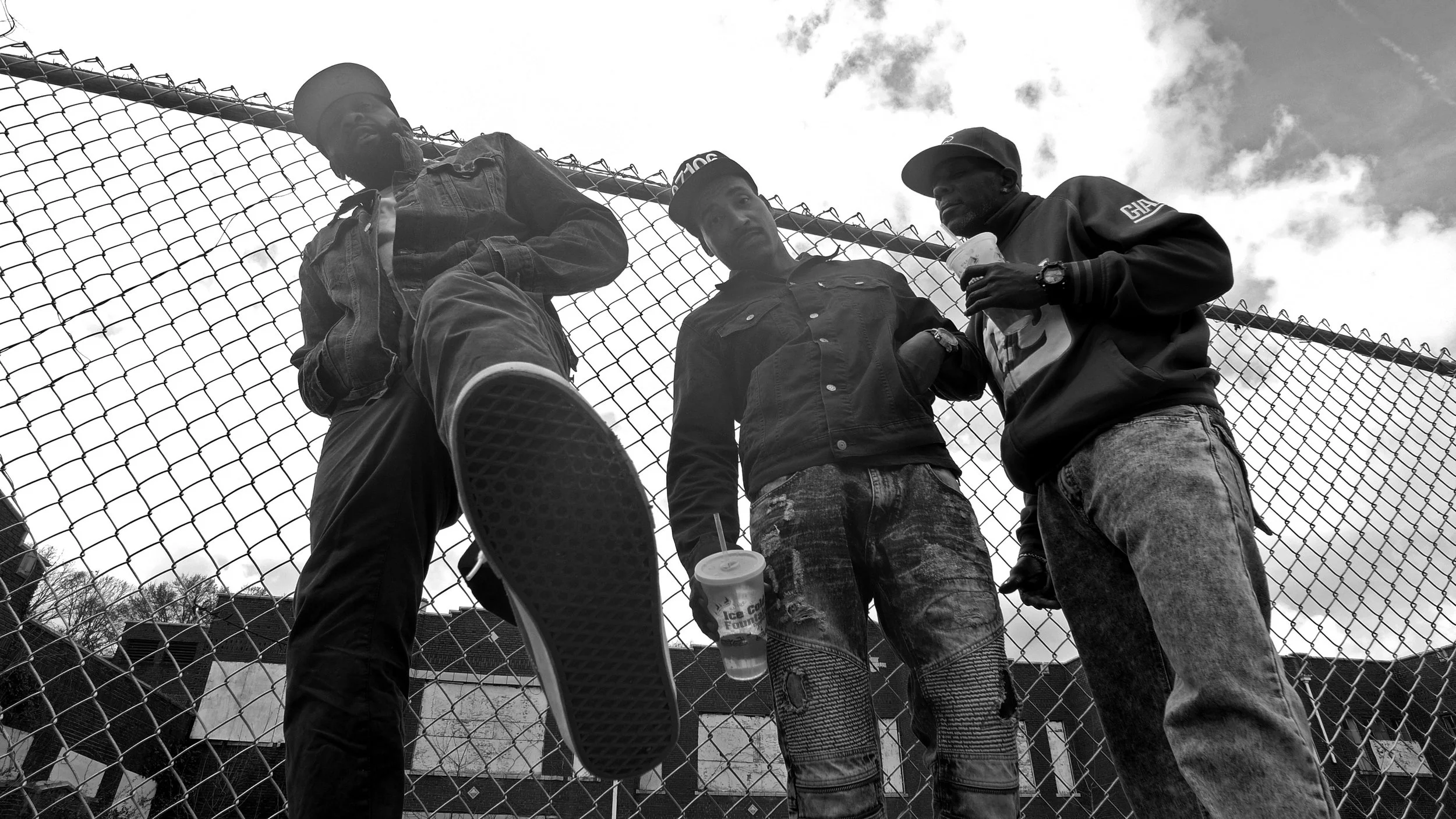 Three young men standing behind a chain-link fence, looking down at the camera, with clouds in the sky overhead.