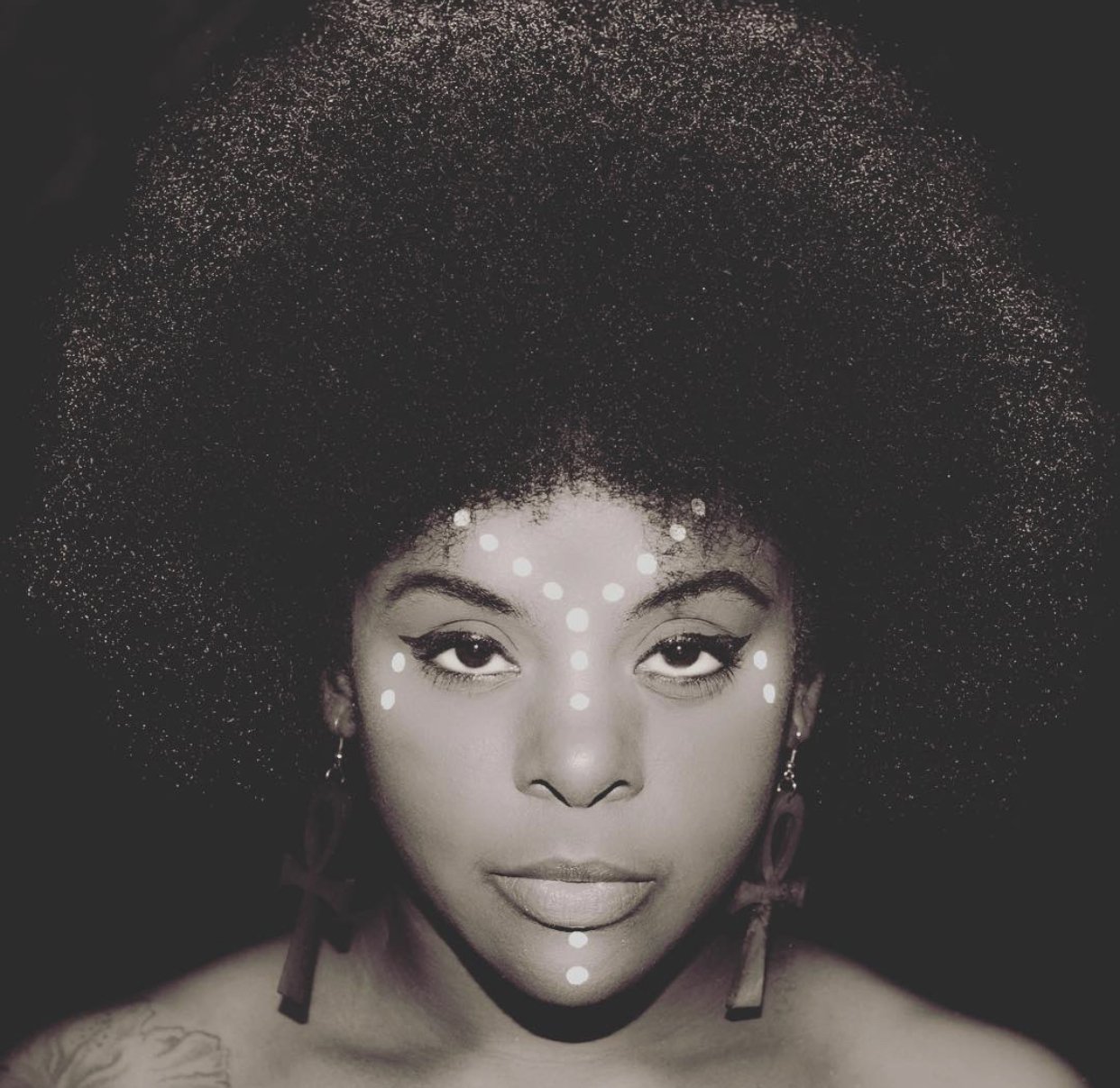 Black and white portrait of a woman with traditional face paint consisting of white dots on her forehead, nose, chin, and cheeks, and an elaborate hairstyle full of natural hair. She has earrings and a tattoo on her shoulder.