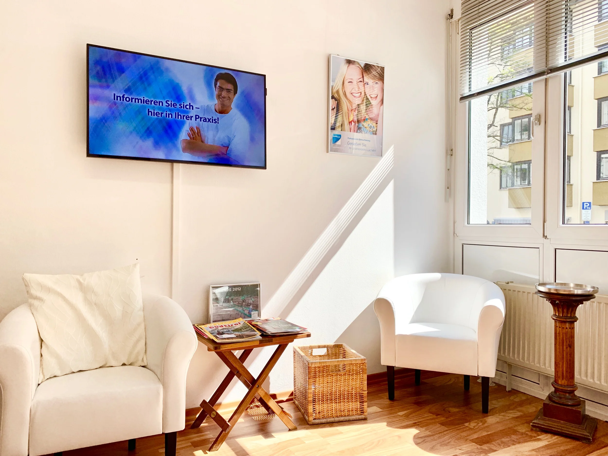 Wartezimmer mit zwei weißen Sesseln und einem kleinen Beistelltisch mit Zeitschriften. Ein Bildschirm an der Wand zeigt medizinische Informationen. Große Fenster lassen viel Tageslicht herein. Ein Plakat mit lachenden Menschen hängt an der Wand.