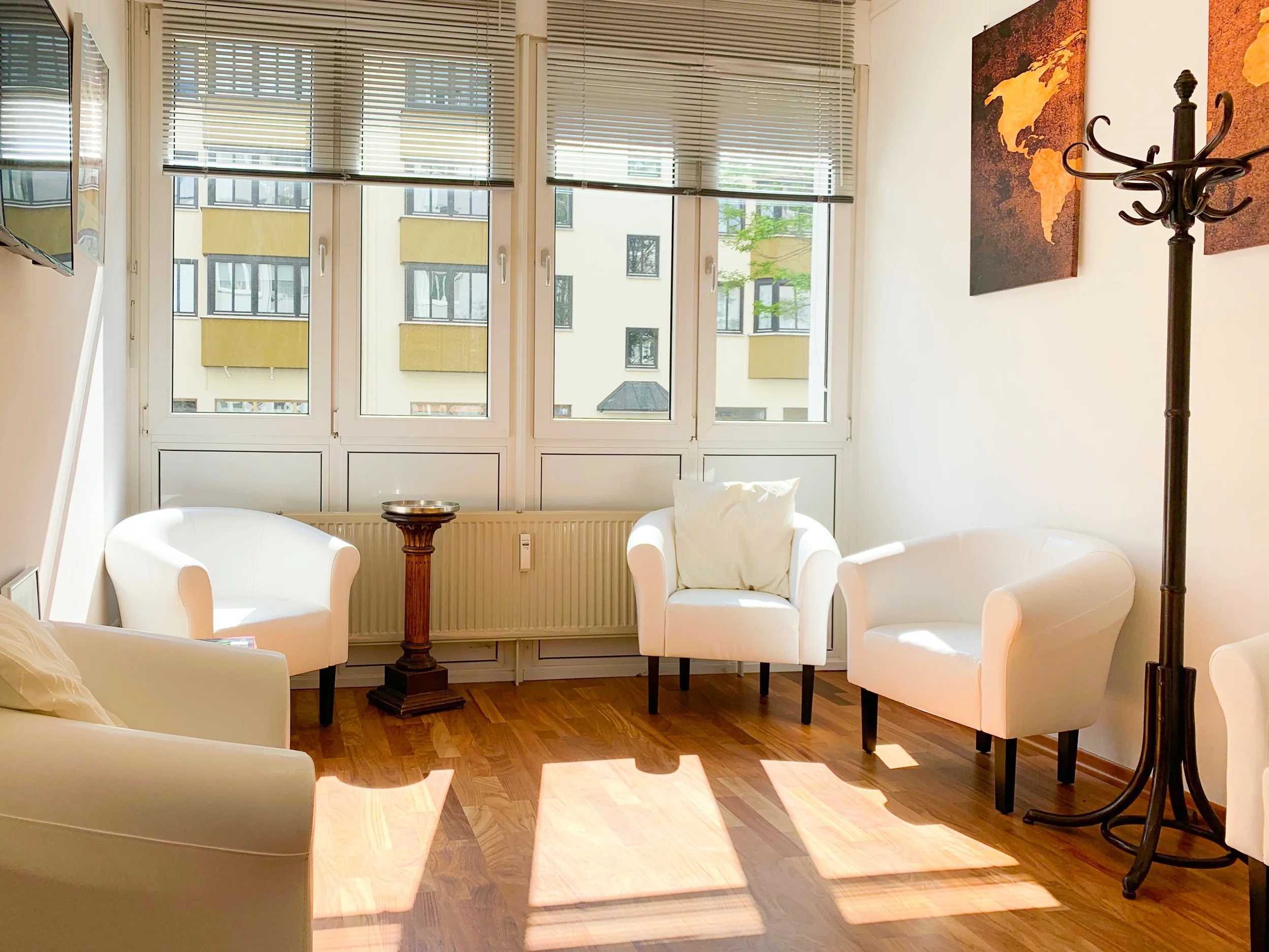 Heller Warteraum mit weißen Sesseln, Fenster mit Jalousien, Wandbildern und Standgarderobe.