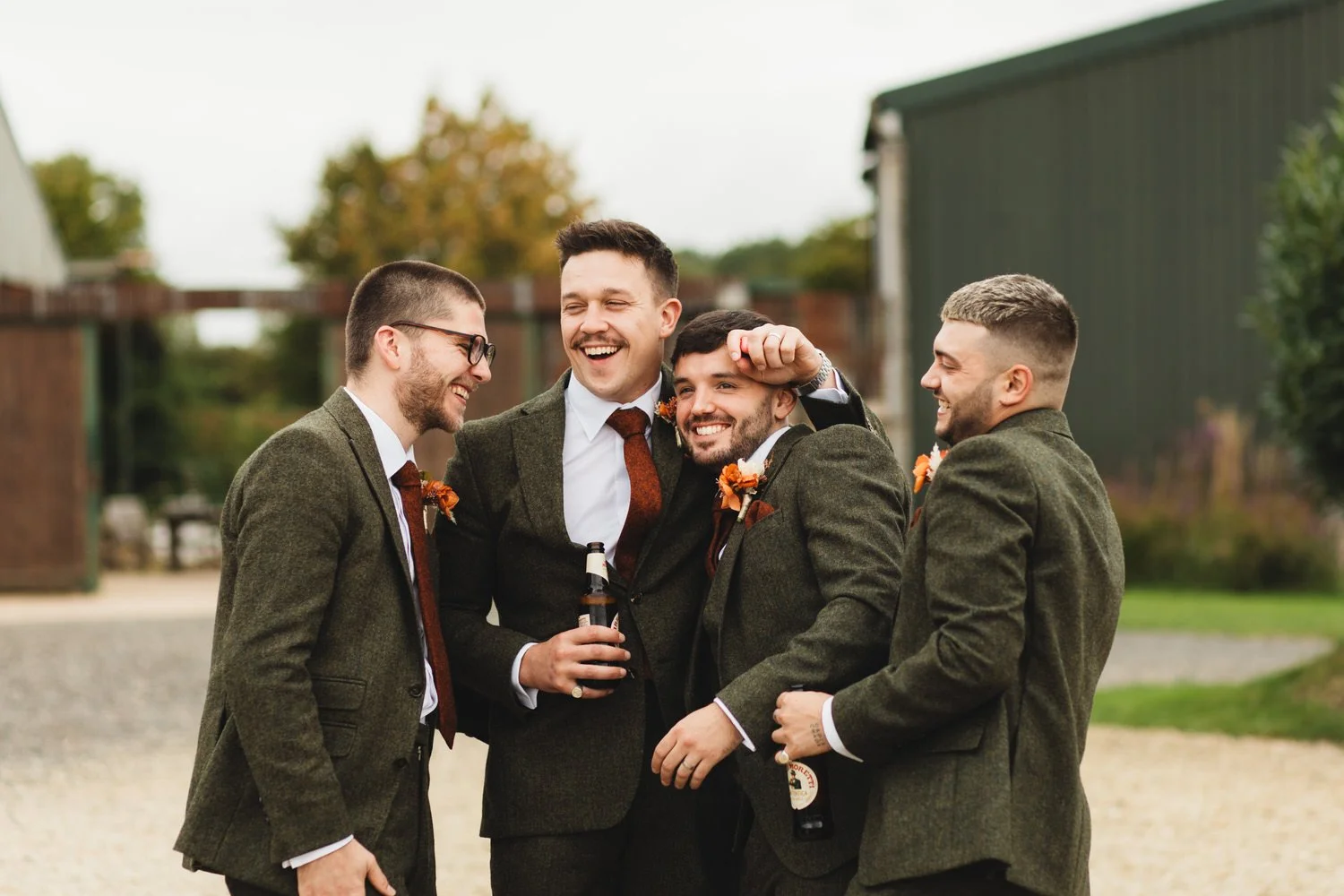 The groom laughs as his friends embrace him. They wear tweed suits with burnt orange ties at Huntsmill Farm.