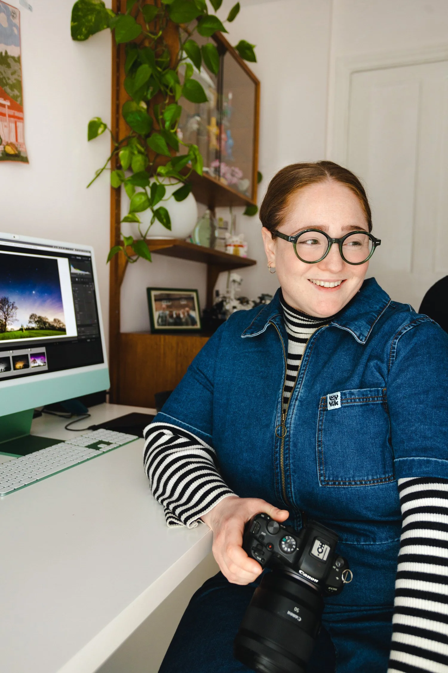 Cat Lane smiles holding her camera in her home office. A computer, framed photo, and shelves with plants are visible in the background.