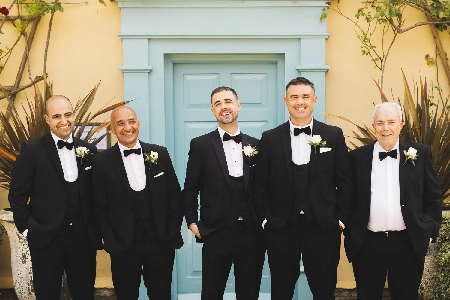 The groom stands in front of a blue door with his four groomsmen. They wear tuxedos and bow ties and laugh together, their hands in their pockets.