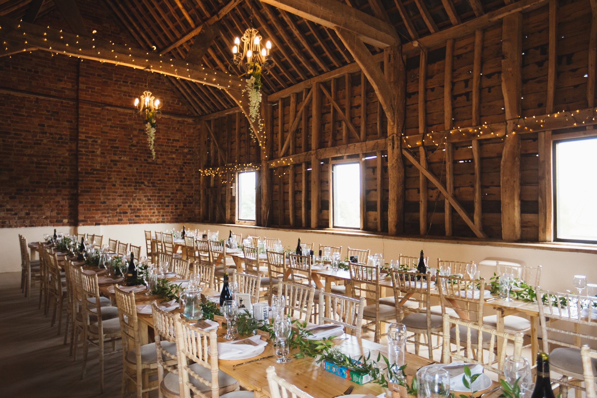 The barn at Newton Park Farm set up for a wedding breakfast. Fairy lights hang from the beams. Wooden tables run along the barn with chiavari chairs and greenery running down the table.