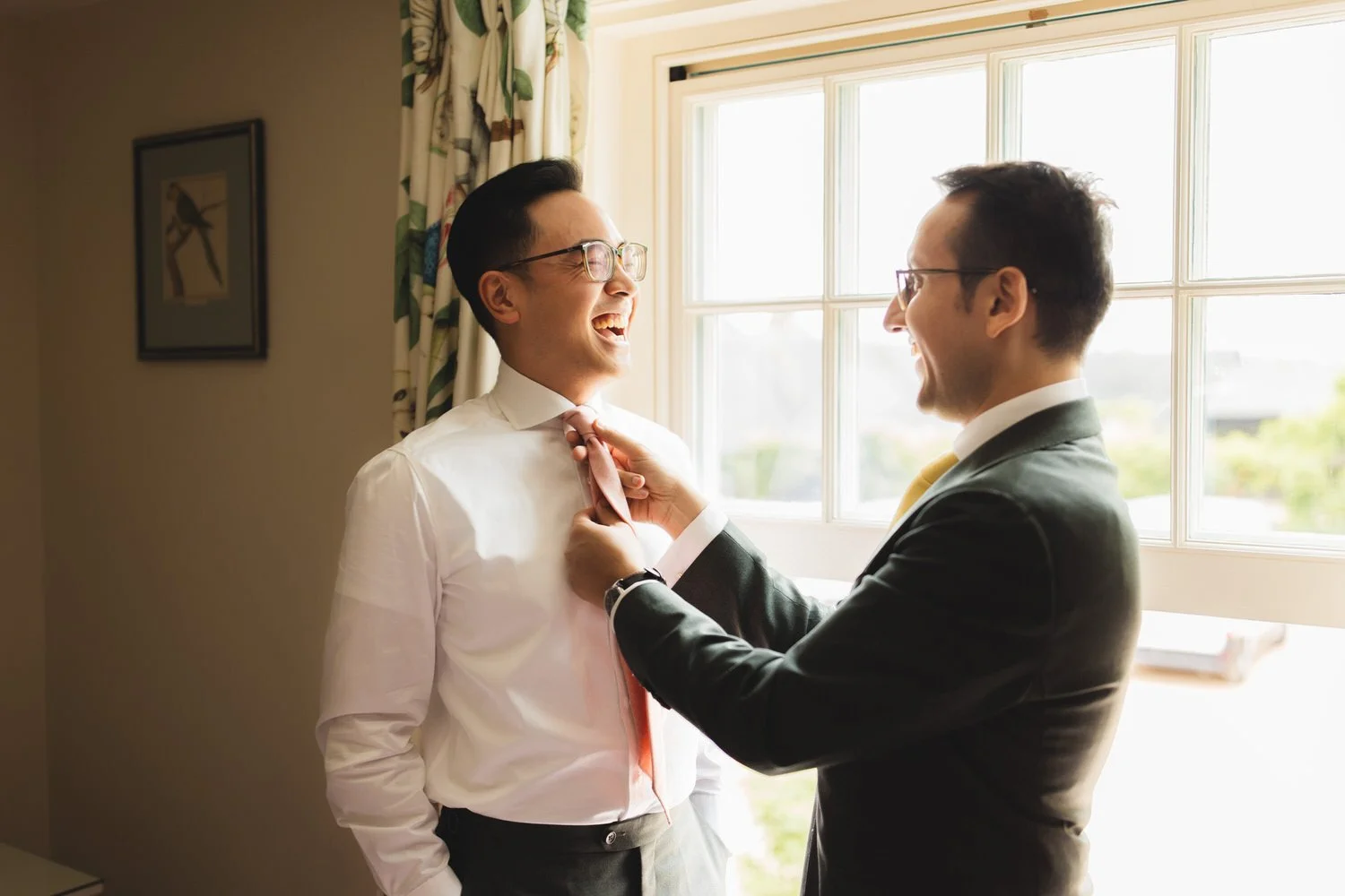 In the Parrot Room at South Farm, the best man helps the groom with his tie in front of the sash windows.