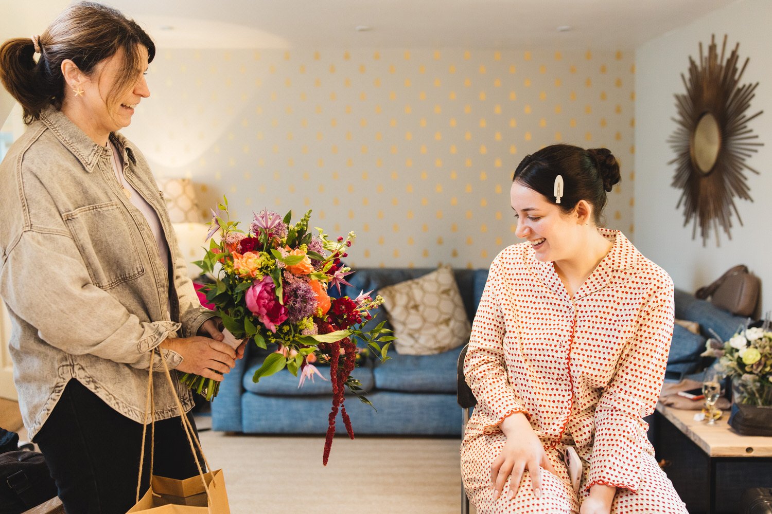In the Coach House Suite at South Farm, a bride in polkadot pyjamas smiles as her florist presents her wedding bouquet to her for the first time.