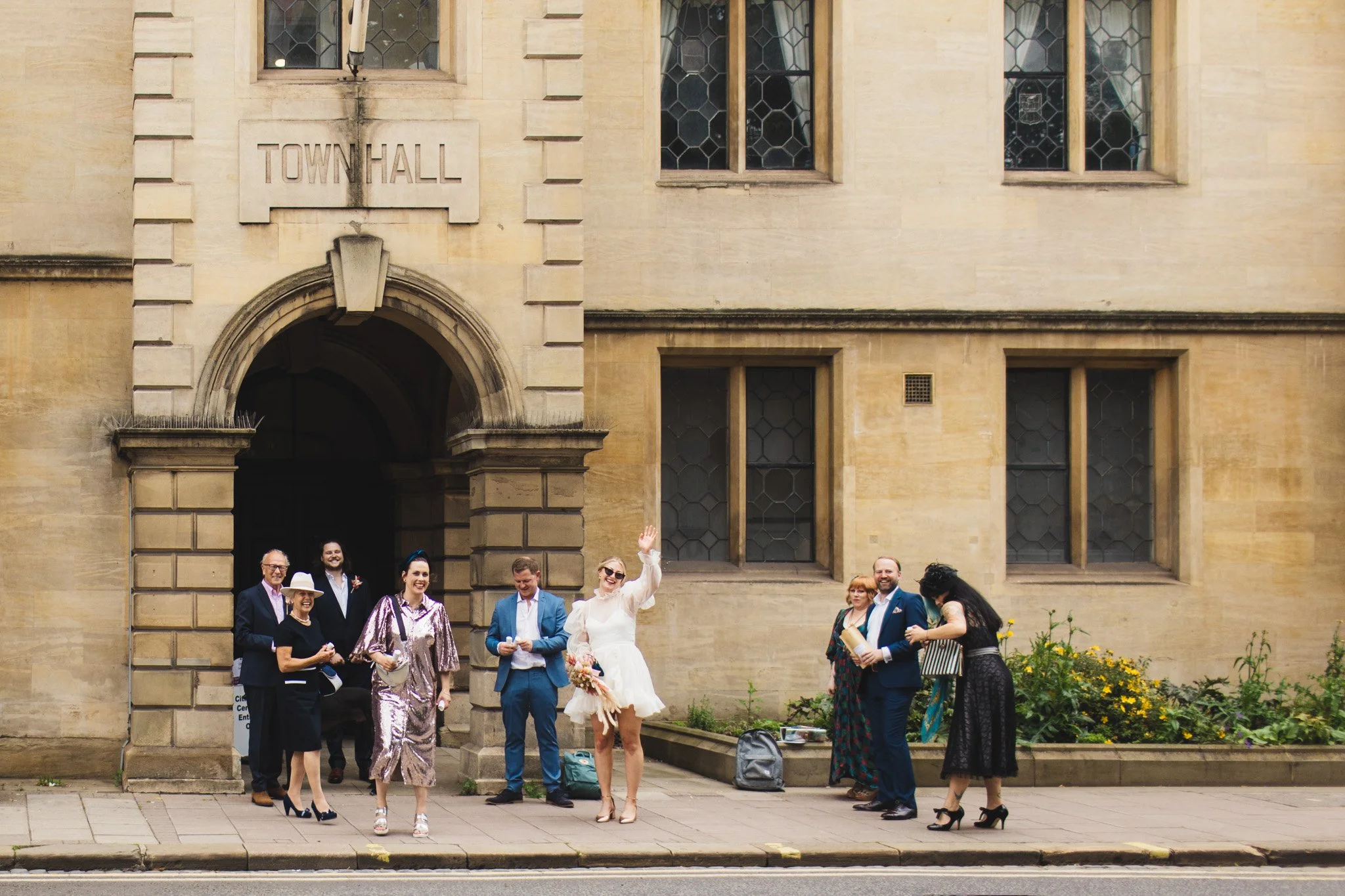 After their wedding ceremony at Bedford Town Hall, the bride waves at the photographer from across the road as the guests laugh.