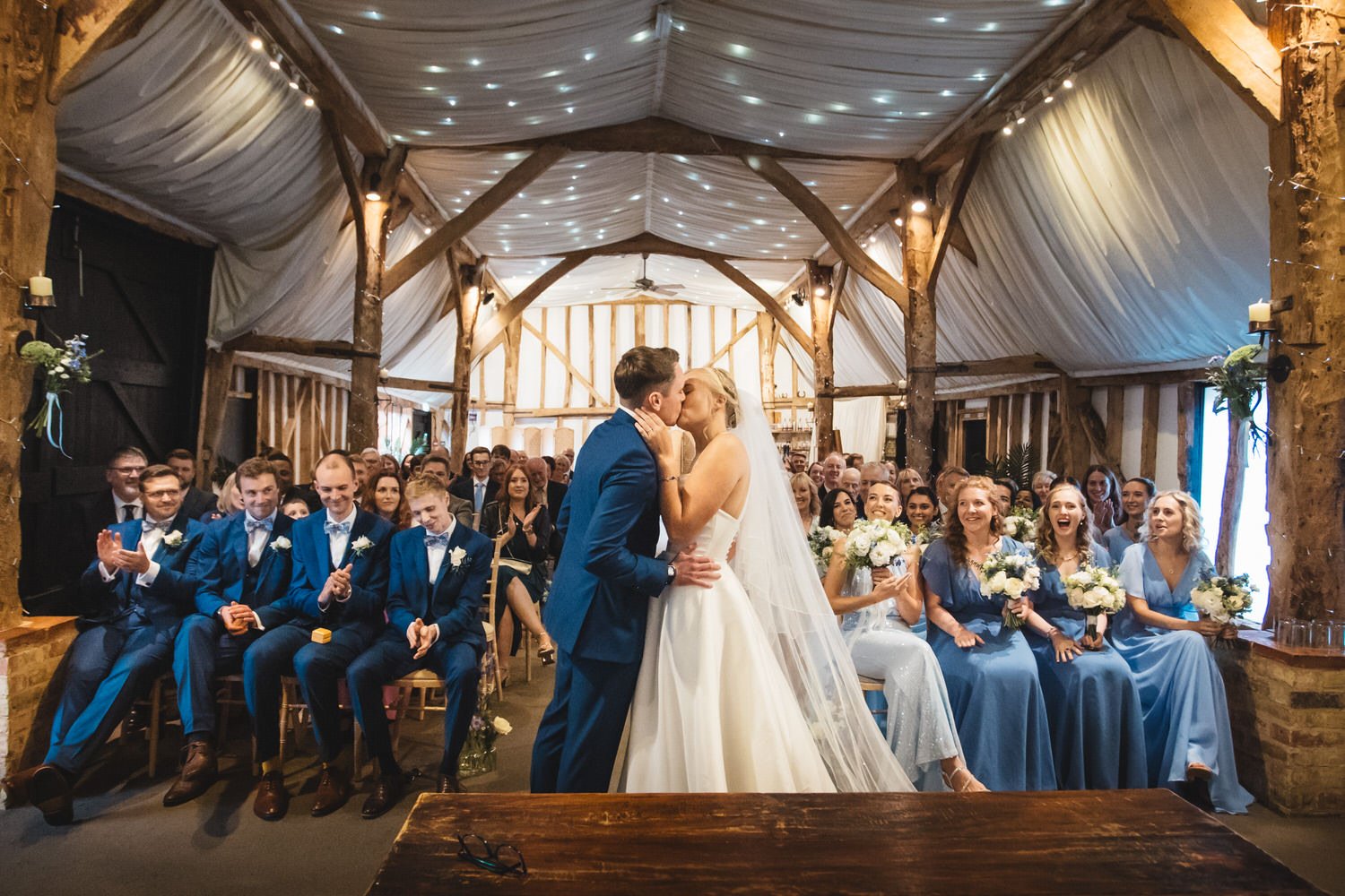 Under the fairy lights in the ceiling of the Tudor Barn at South Farm, a couple kiss at the end of their wedding ceremony. Their guests cheer and applaud in the background.