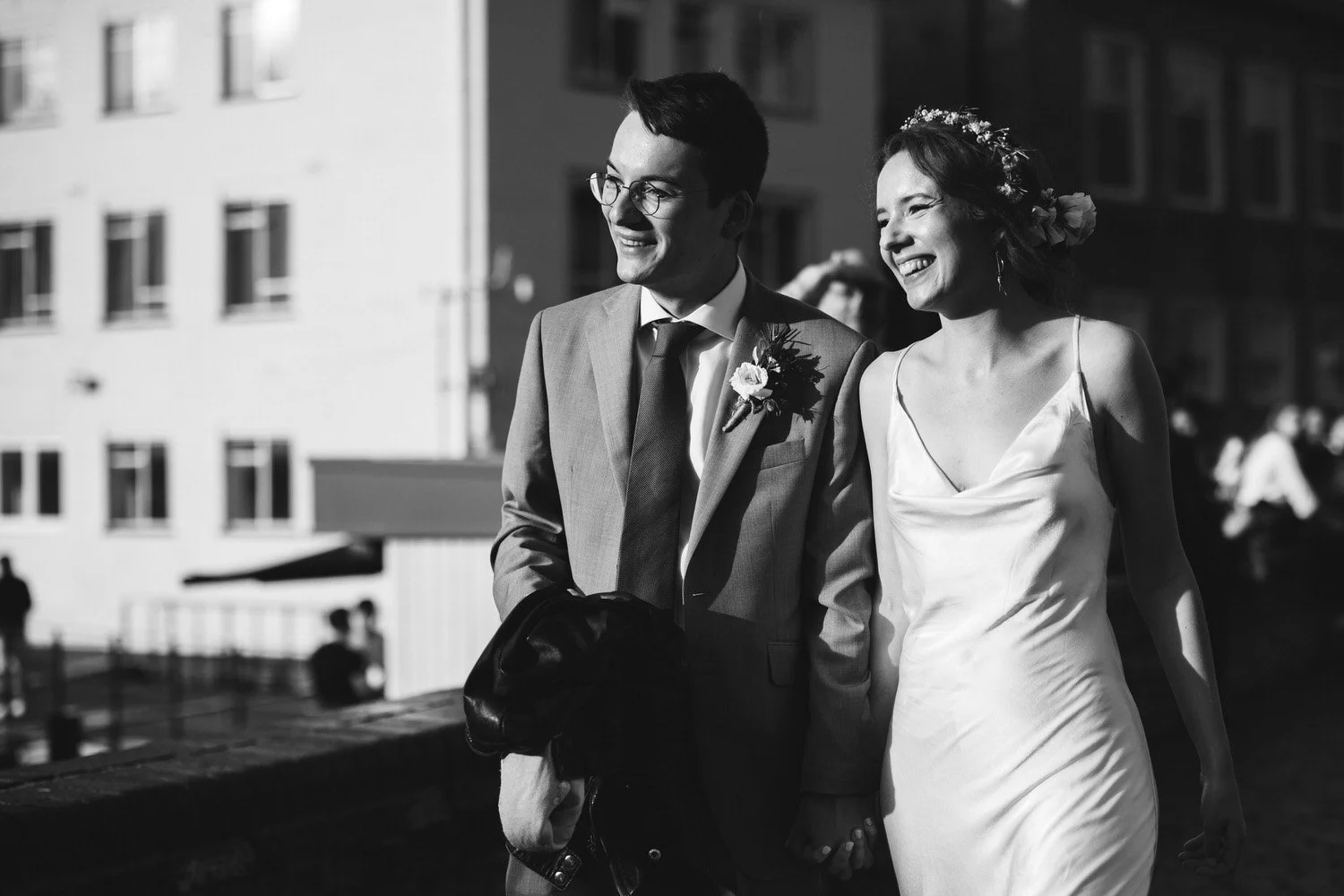 A couple smile with the evening sun on their faces as they walk through Cambridge city centre at the end of their wedding day. The bride wears a flower crown and satin dress.