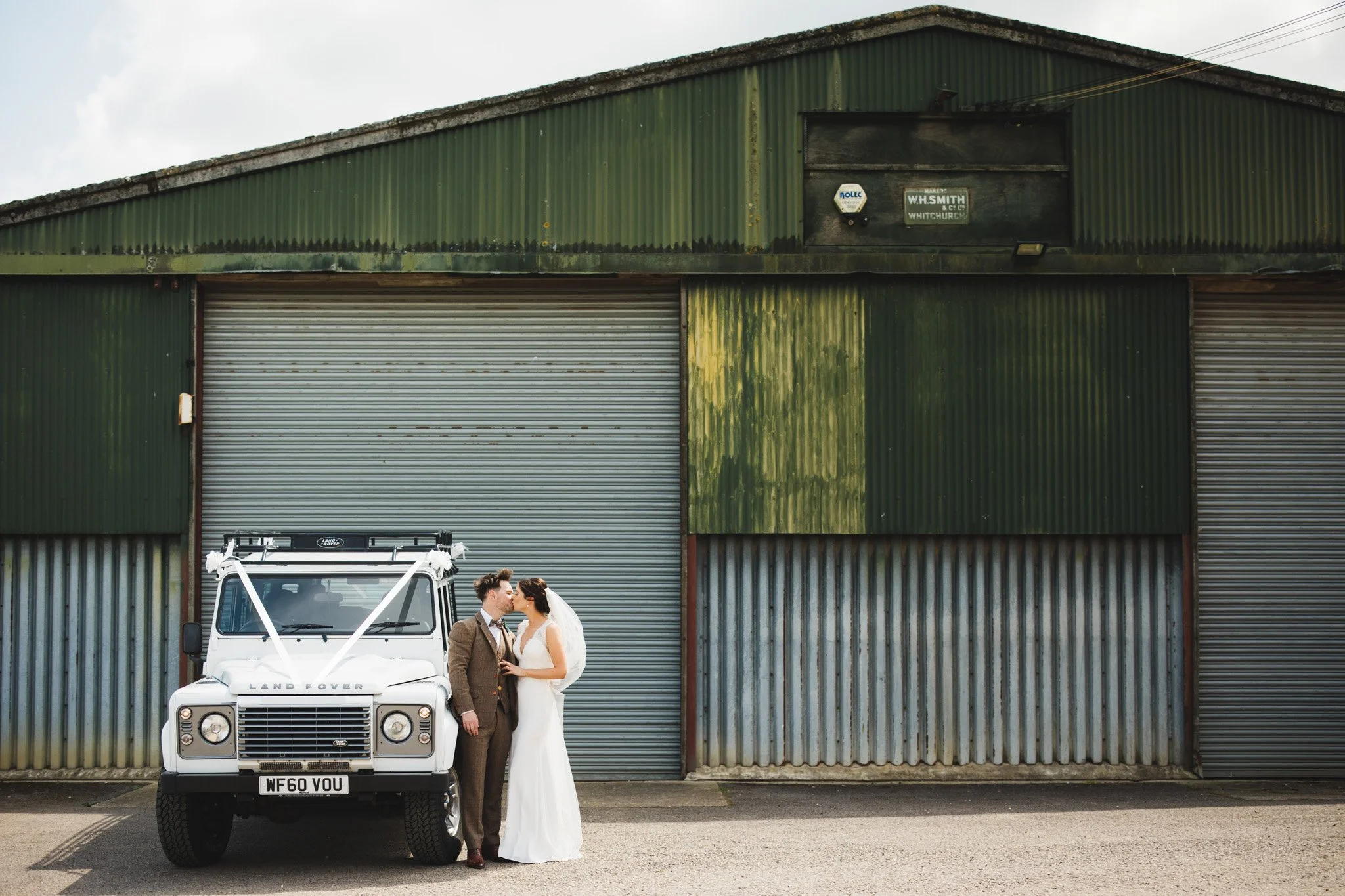 Newlyweds share a kiss next to their Land Rover wedding car, in front of a corrugated steel barn.