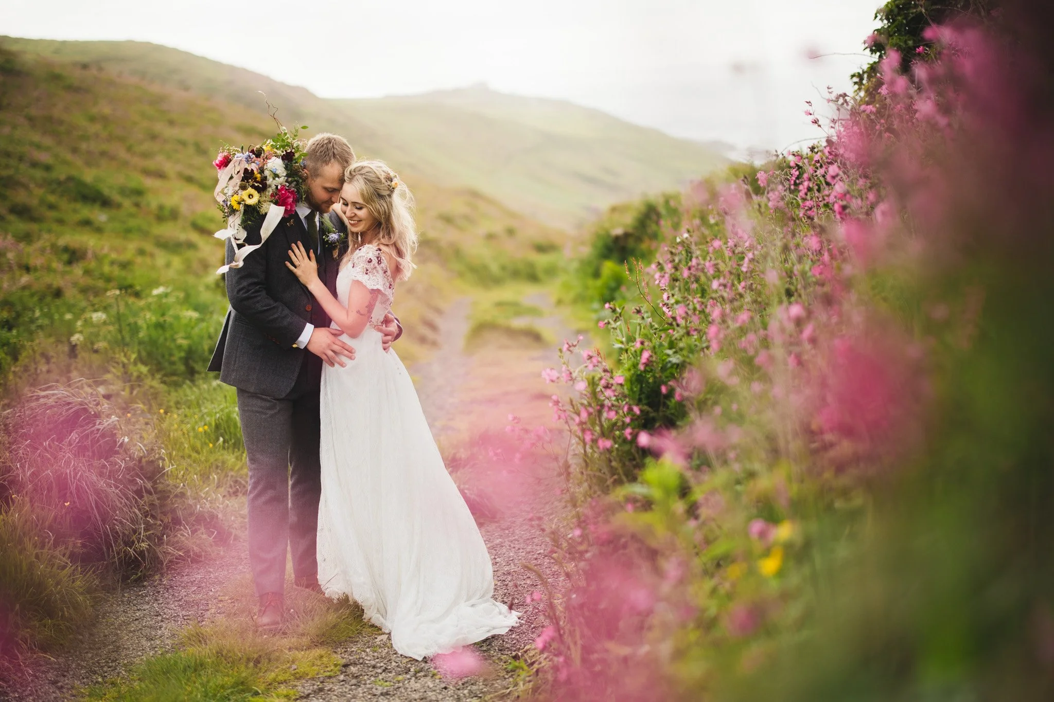 A bride and groom embrace on a scenic nature trail with pink flowers in the foreground, green hills, and cloudy sky in the background on their seaside wedding day.