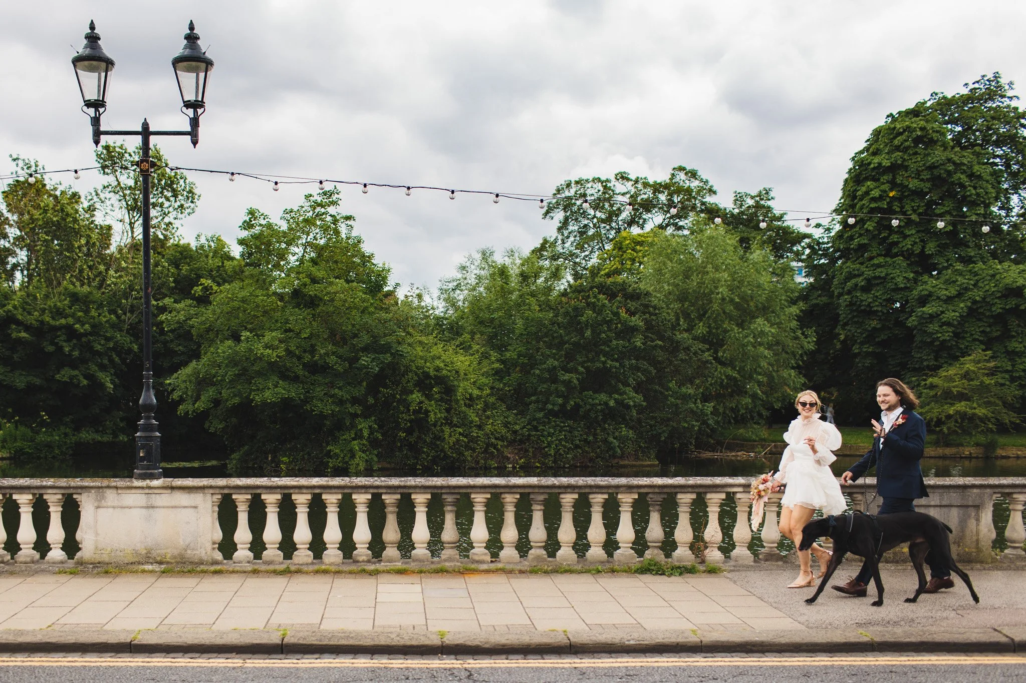 A bride and groom walk down the Embankment in Bedford on their wedding day with their dog. The river and festoon lights are in the background.
