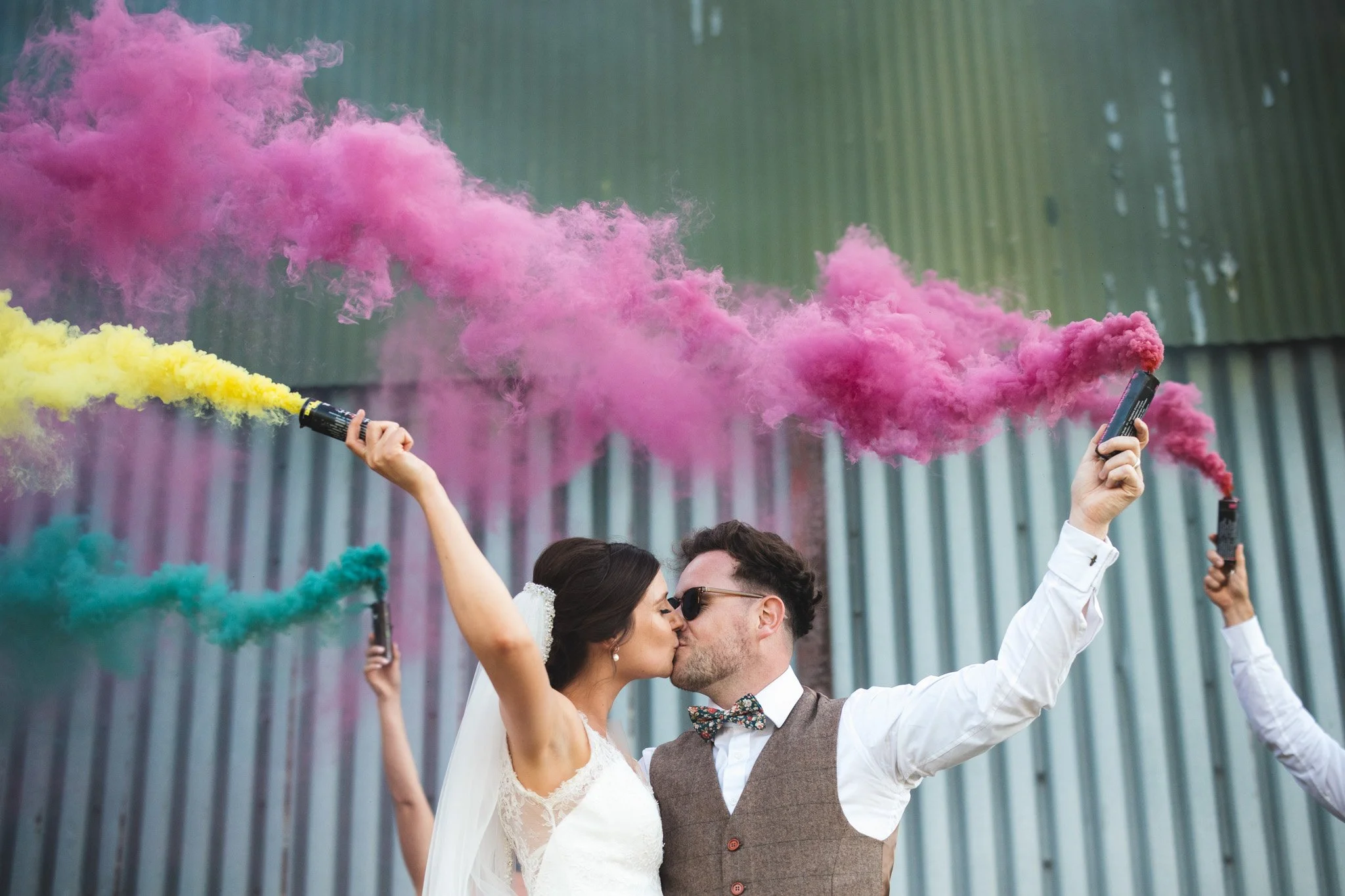 Alex and Matt share a kiss as they hold smoke bombs, vibrant plumes of red and yellow smoke in the air above them.