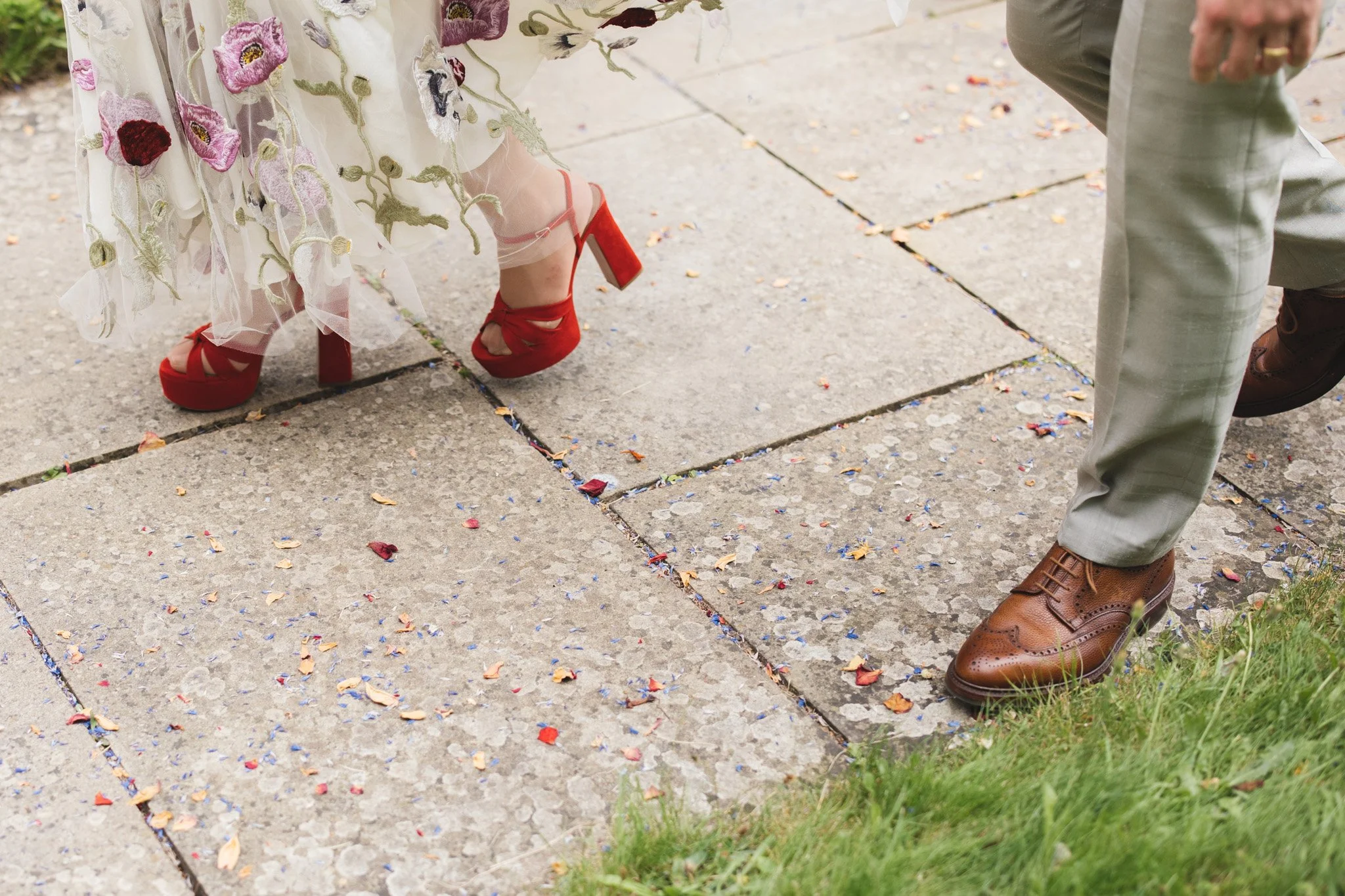 A detail shot of the couples feet as they walk down the church path. She wears a dress embroidered with flowers and red platform heels. The groom wears a tan suit with checkered pattern and brown leather brogues.