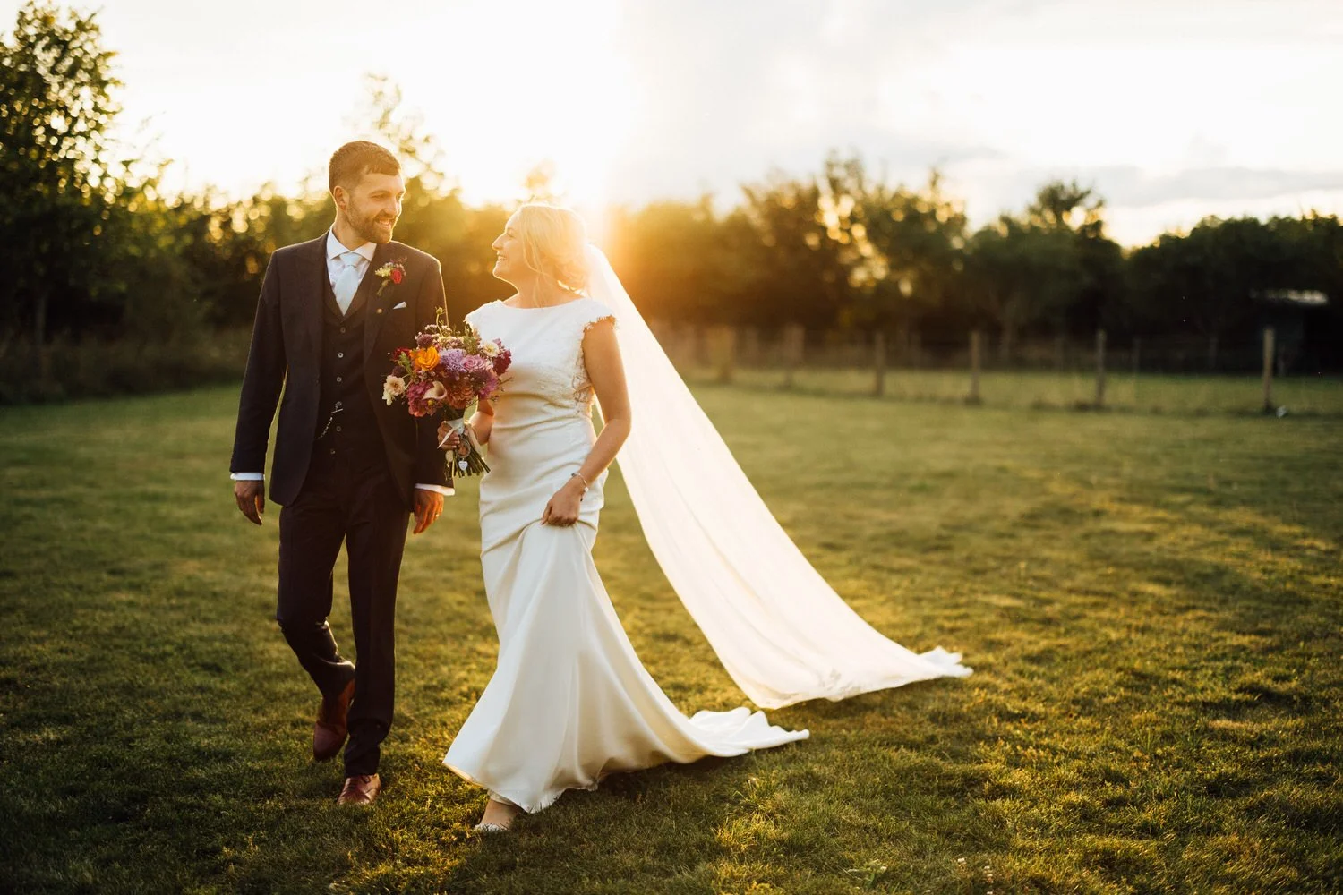A couple walk in the fields at South Farm as the evening sun sets behind them. The bride's veil flows out behind her.