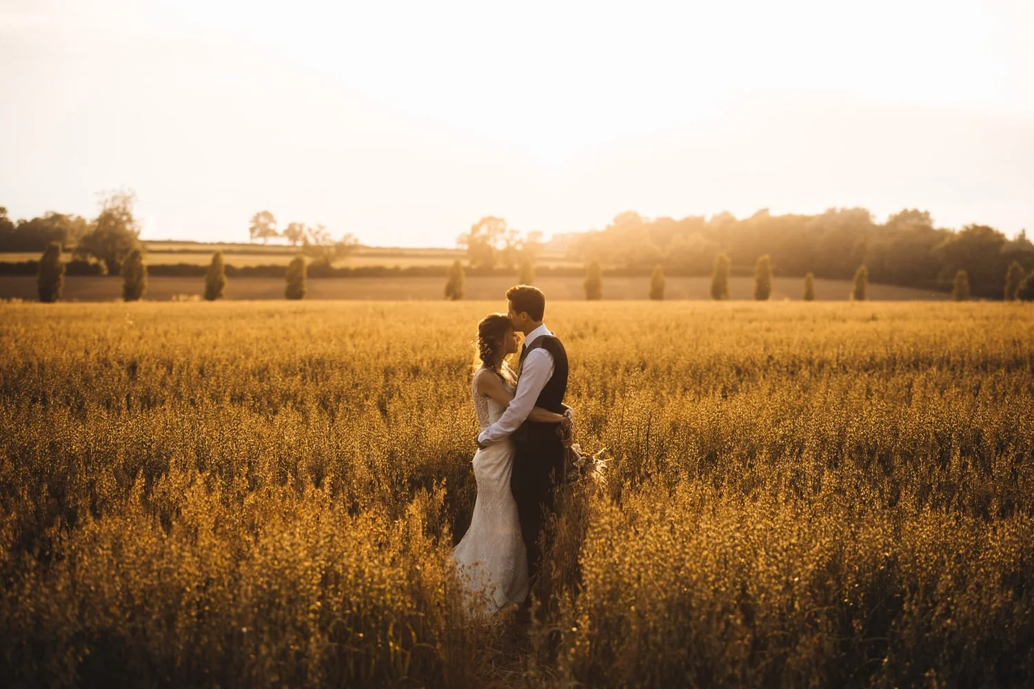 A couple dressed in wedding attire embracing in a golden field during sunset at Huntsmill Farm.