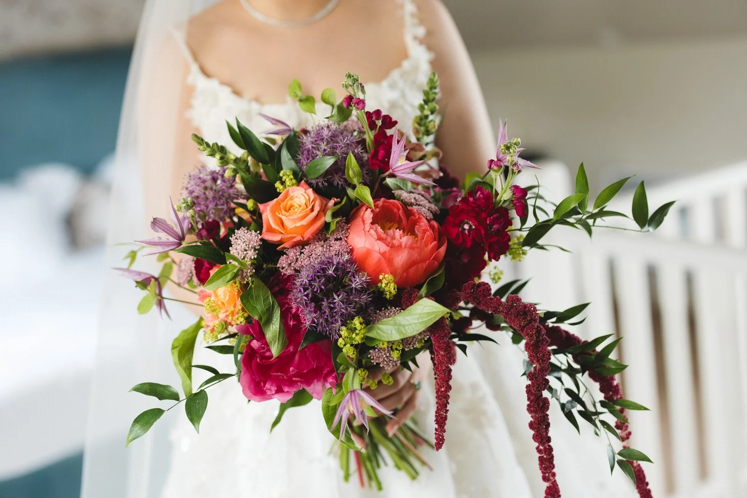 A close up of a vibrant bouquet of flowers held by a bride. Orange roses, pink peonies and purple alliums with trailing ruscus.