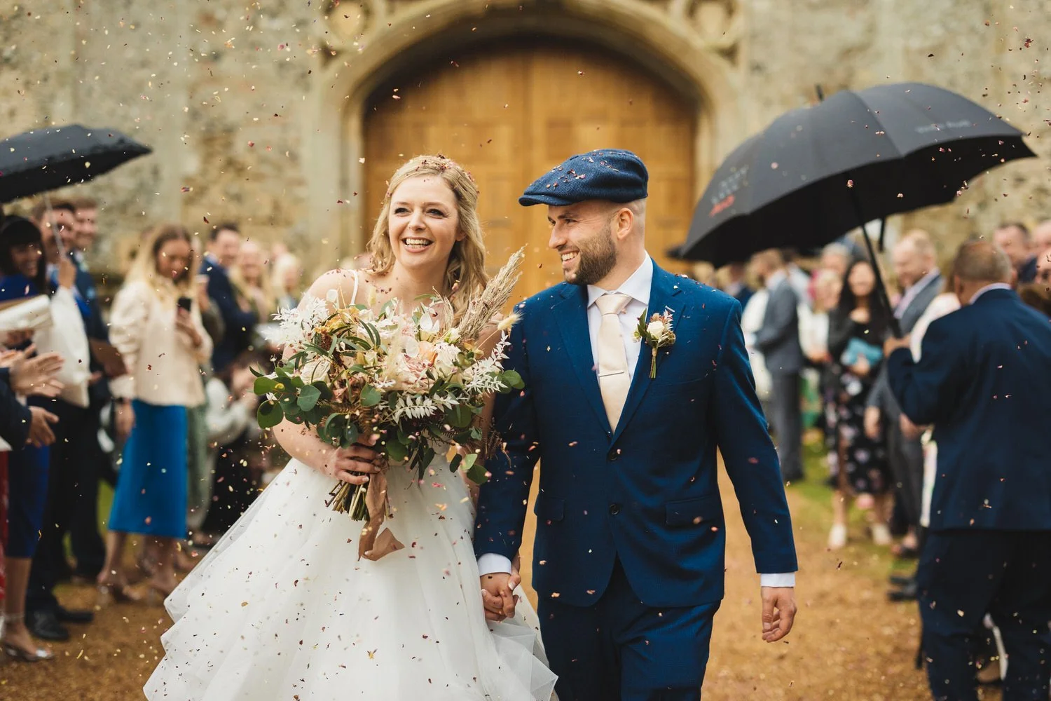 On a rainy day, a bride and groom smile as they walk through their confetti moment at Pentney Abbey in Norfolk. Guests carry umbrellas. The bride has blonde hair and carries a bouquet of dried flowers, and the groom wears a blue suit and matching fla