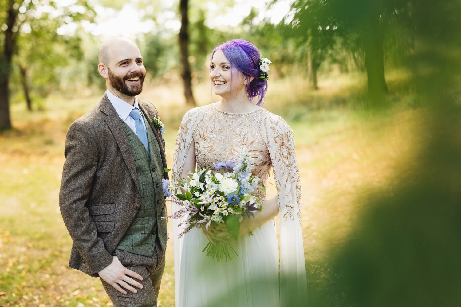 The groom in a tweed suit laughs off to the side as his bride laughs with him. She has purple hair and wears a dress with gold and bronze embroidery and carries a bouquet of wildflowers.