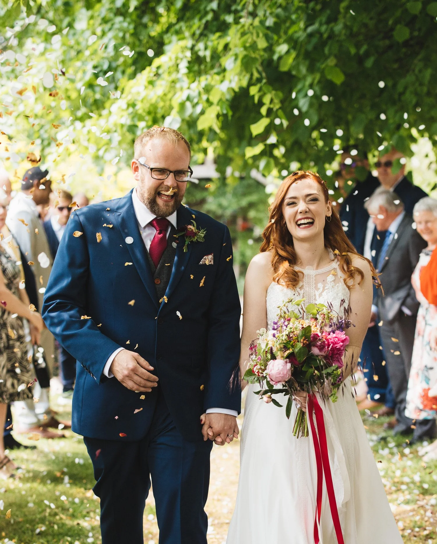 A happy bride and groom holding hands and smiling at their wedding outdoors, surrounded by guests and green trees, with confetti falling.