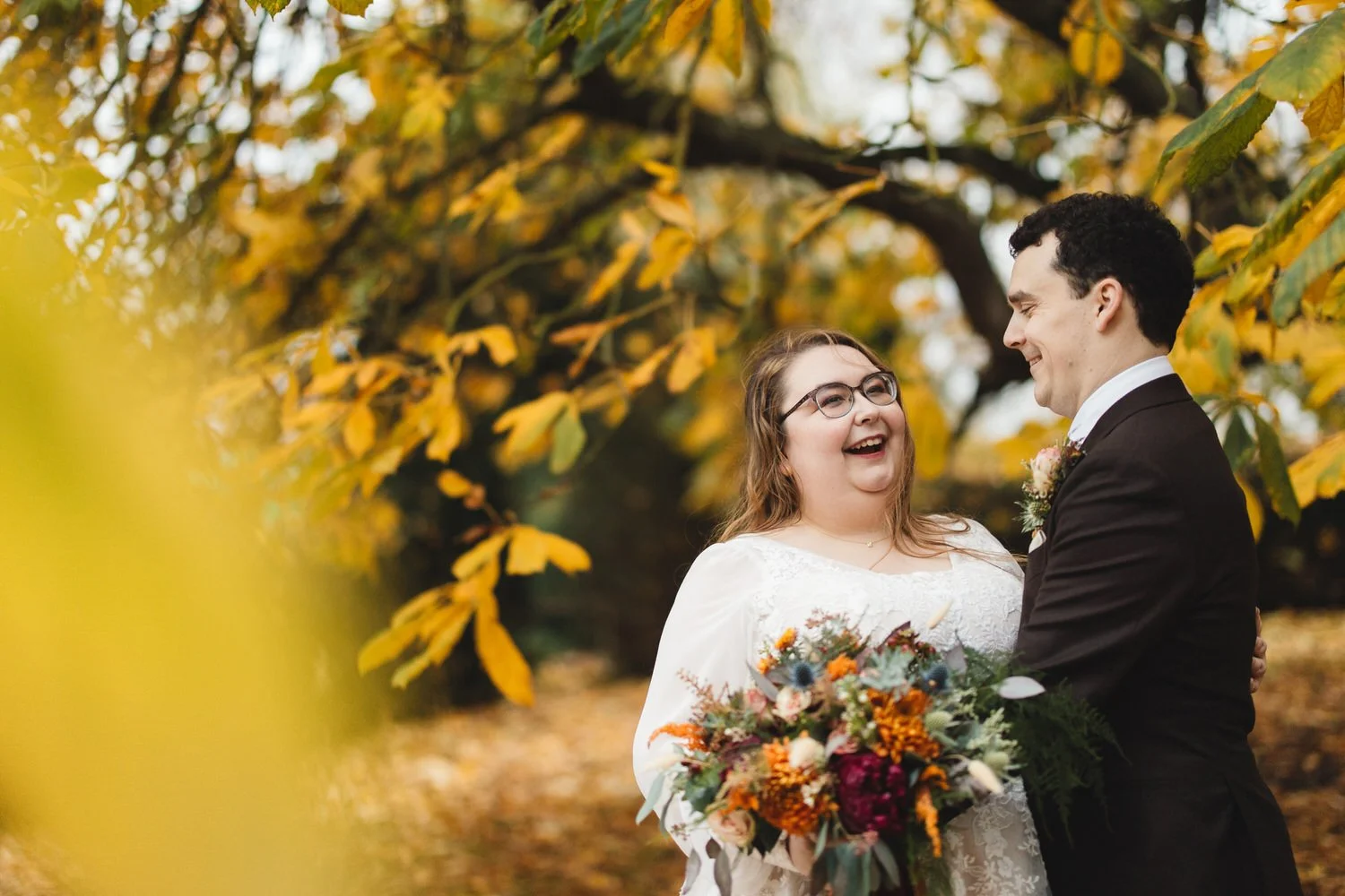 Autumn leaves surround a couple as they laugh on their wedding day. The bride wears glasses and a white dress, and the groom is in a brown suit. She laughs off to the side as she holds a bouquet of orange and pink flowers.