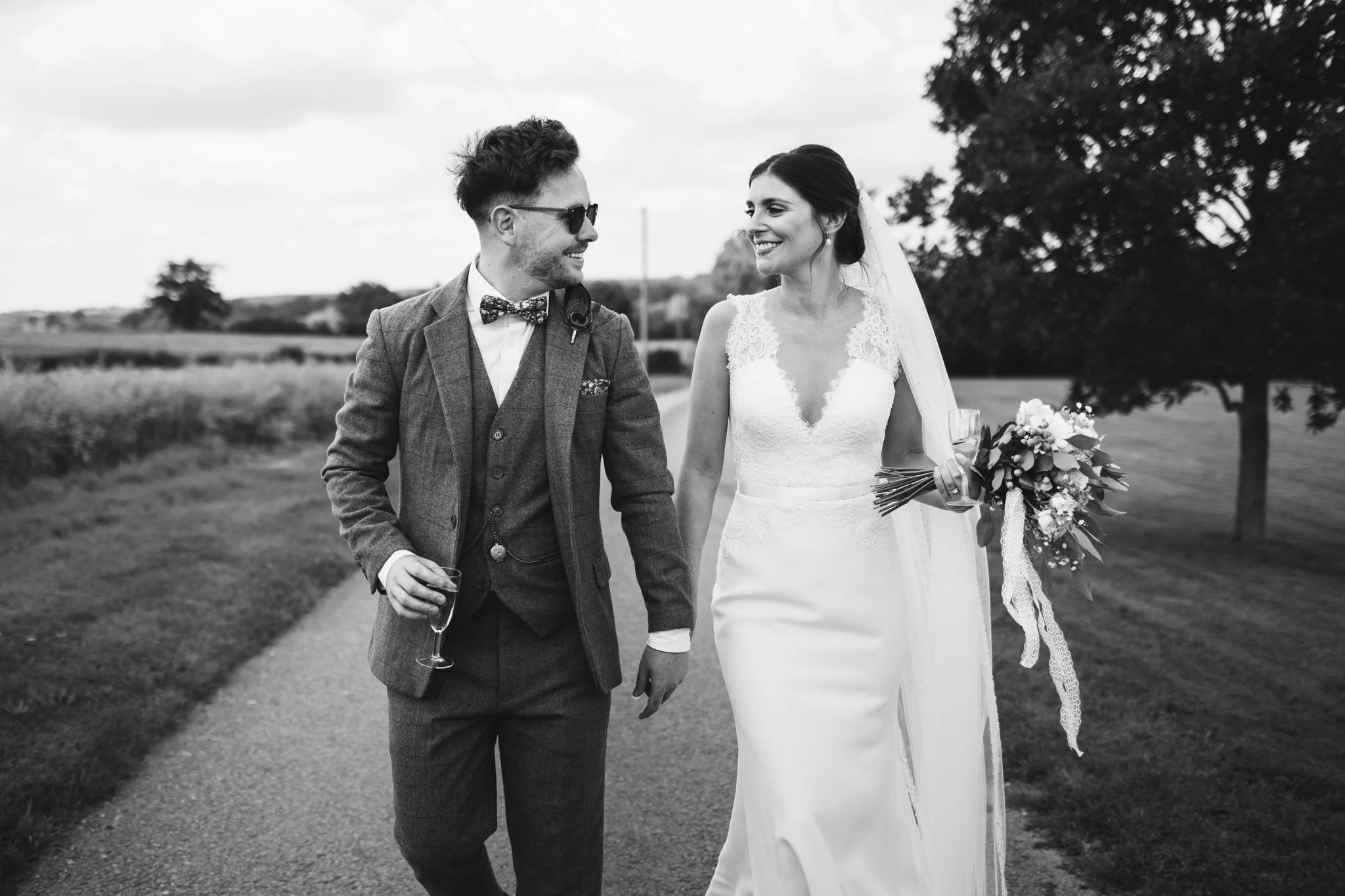 Alex and Matt walk down the driveway at Newton Park Barns. They smile at each other, each carrying a glass of champagne.