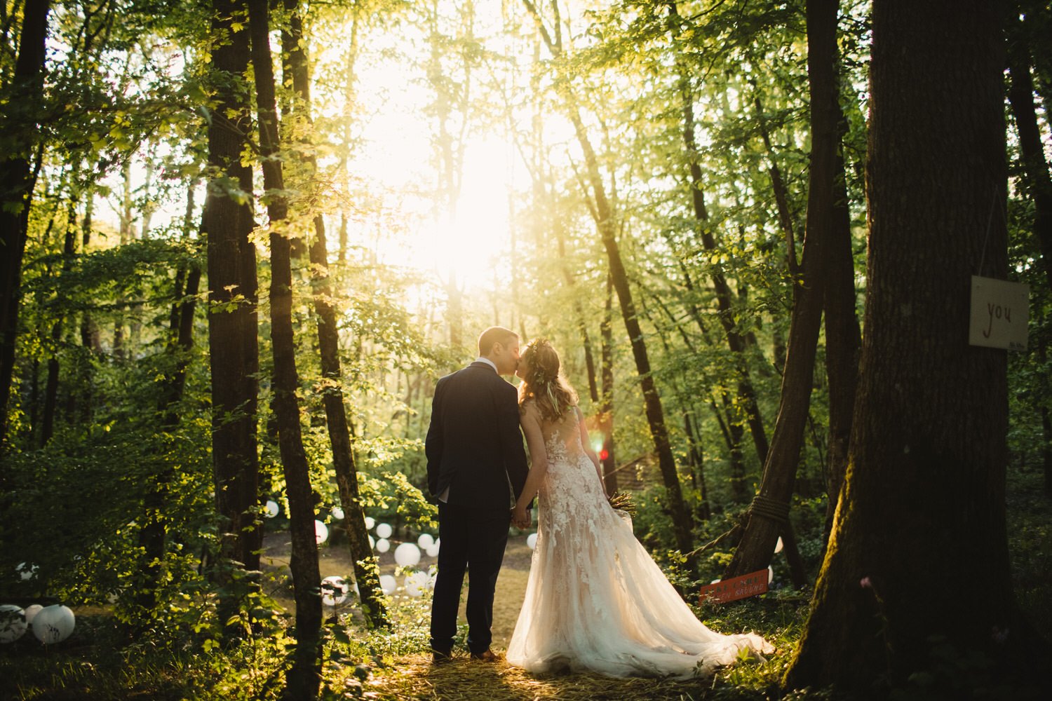 Bride and groom holding hands and kissing in a sunlit forest after their countryside outdoor wedding, with trees and white lanterns on the ground.