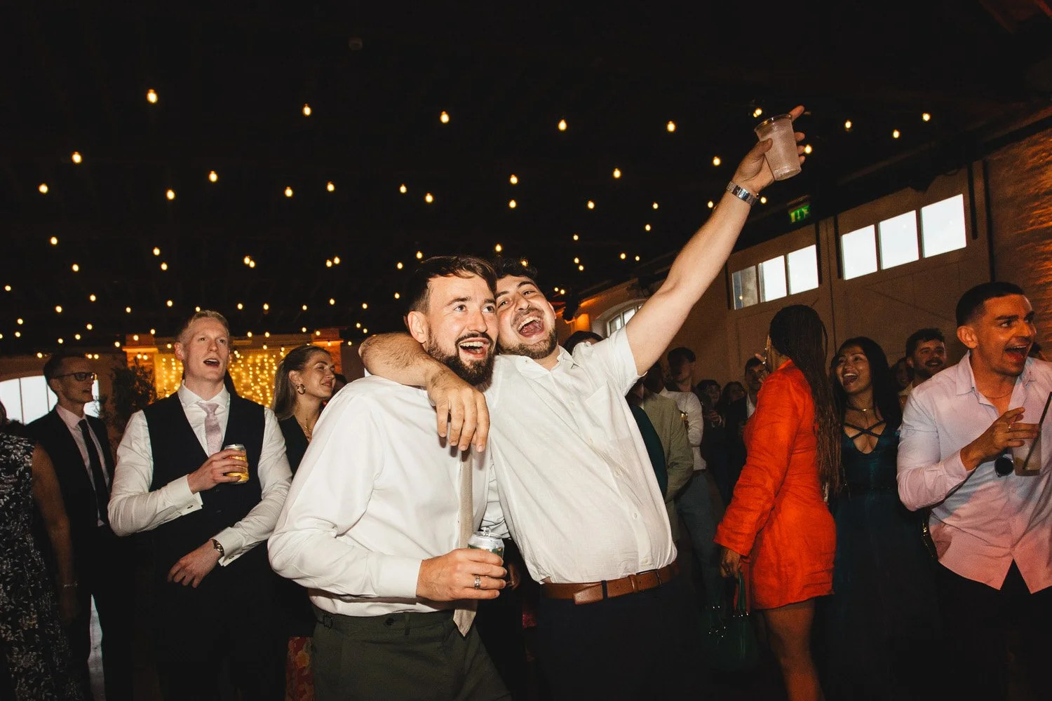 On the dancefloor, a friend wraps his arm around the groom and pumps his fist in the air as they sing to the wedding band with festoon lights in the background.