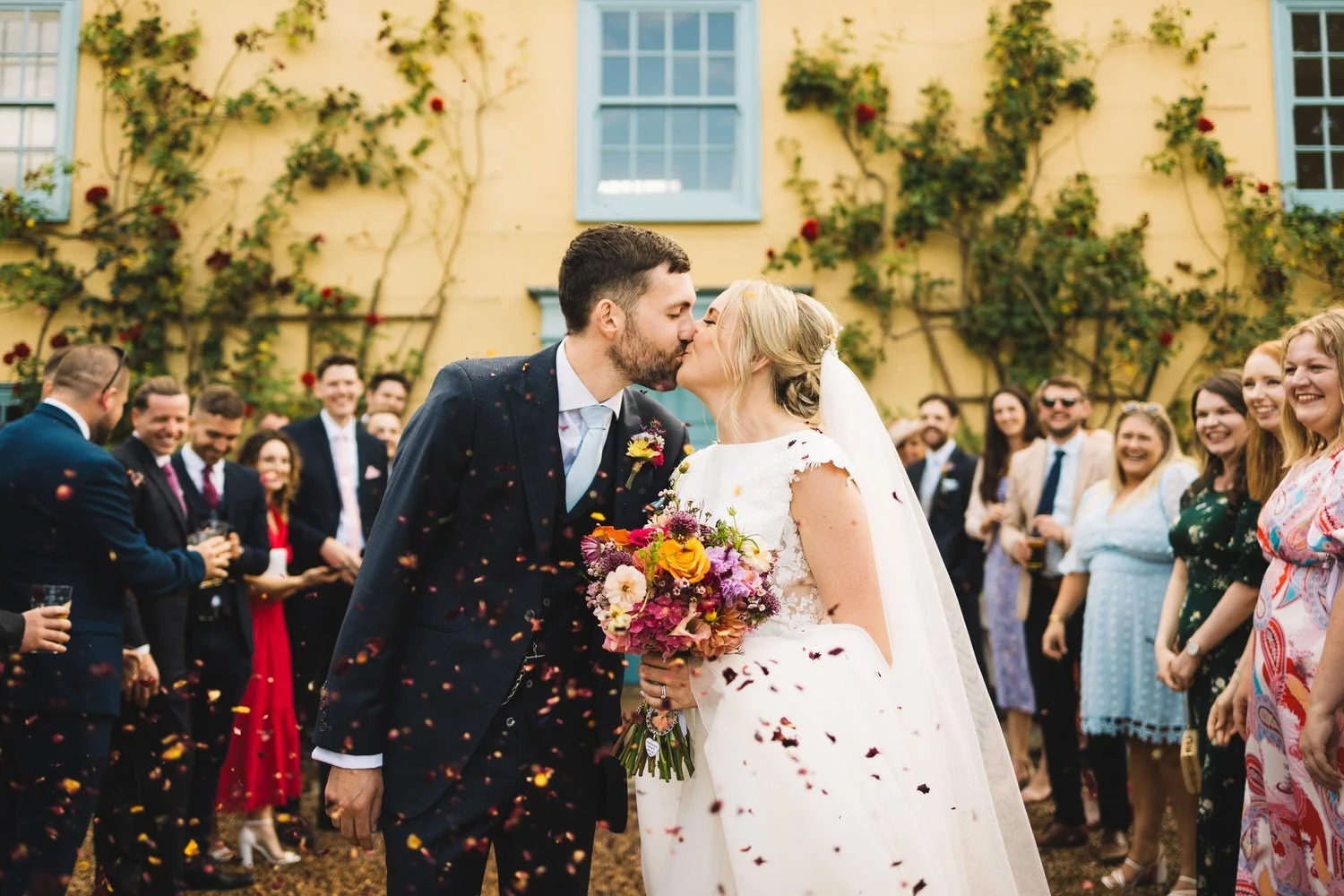 A bride and groom kiss during their wedding celebration while their guests watch and throw confetti in the air at South Farm in Hertfordshire.