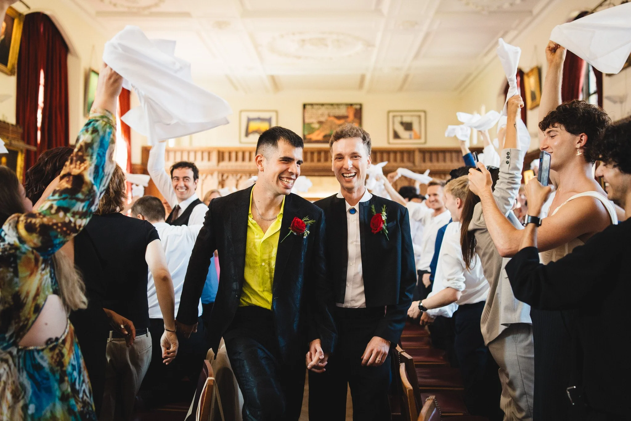 Two grooms smile as they walk into the wedding breakfast between lines of guests cheering and waving their napkins in the air.