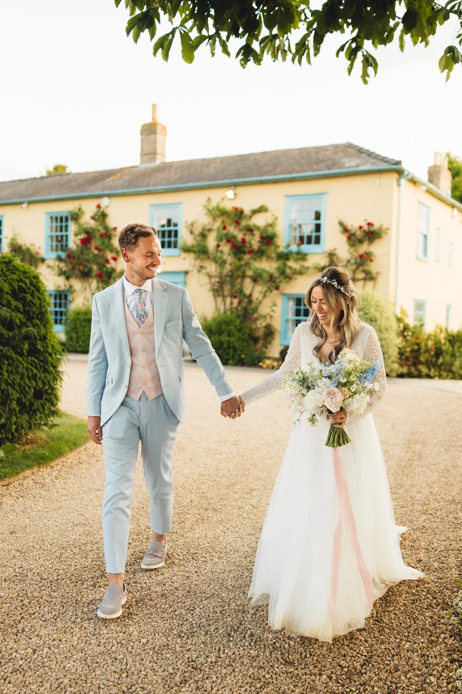 A bride and groom take a walk on their wedding day at South Farm in Hertfordshire in front of the yellow farmhouse with blue windows. The groom wears a light blue suit and the bride carries a bouquet of pastel flowers.