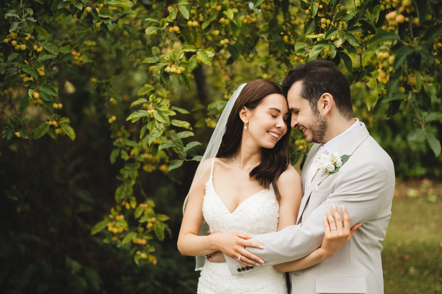 A bride and groom lean their heads together and smile at Huntsmill Farm in Buckinghamshire. They are outdoors with greenery and yellow flowers in the background.