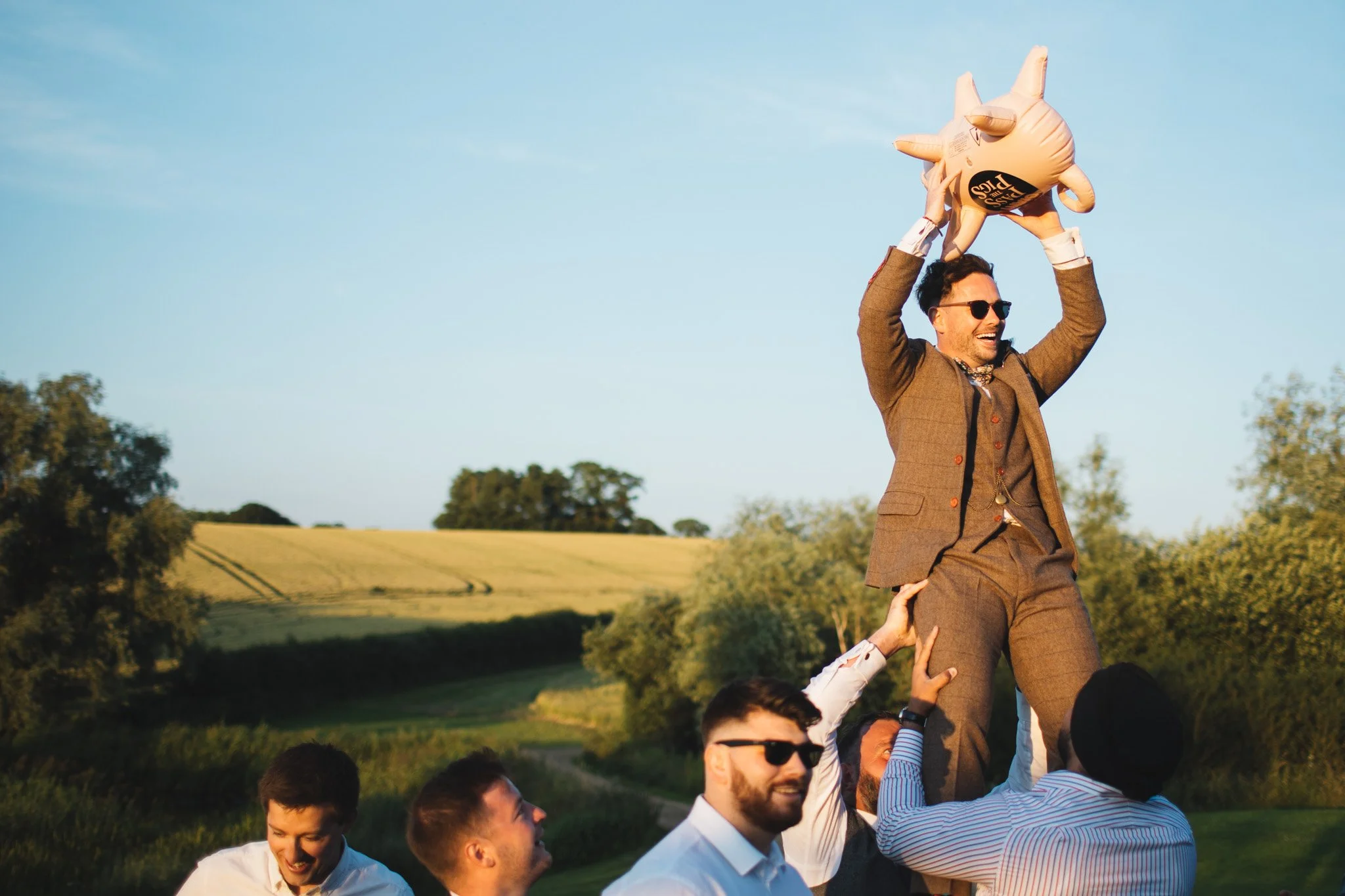 Guests lift the groom up to catch a big inflatable Pass the Pigs. He smiles, sunglasses on, as the evening light hits his face.