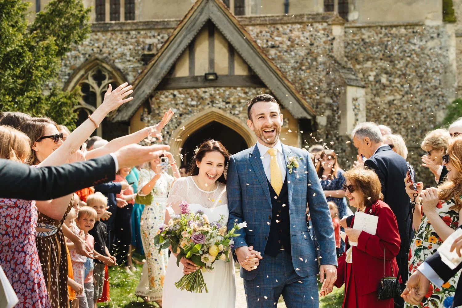 Happy newlywed couple walking hand-in-hand through a crowd of friends and family outside of a Cambridgeshire church, with confetti falling around them.