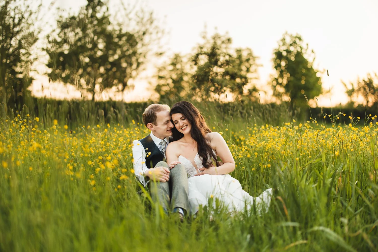 A couple sit in a field of buttercups at South Farm. The sun sets behind them as they cuddle up.